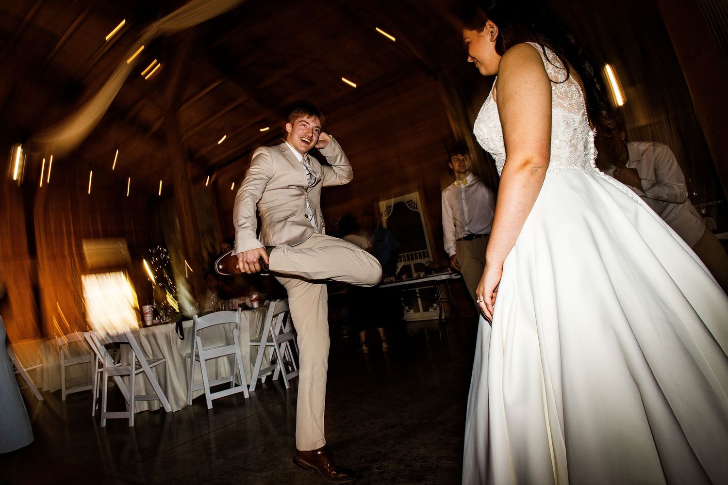 Groom in tan suit dancing energetically with bride in white gown during wedding reception in rustic barn venue.