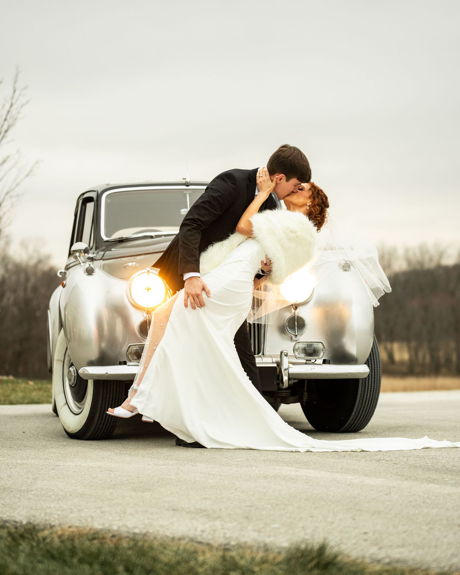 A romantic couple poses in front of a classic silver car in a series of artistic wedding photos on a cloudy day.