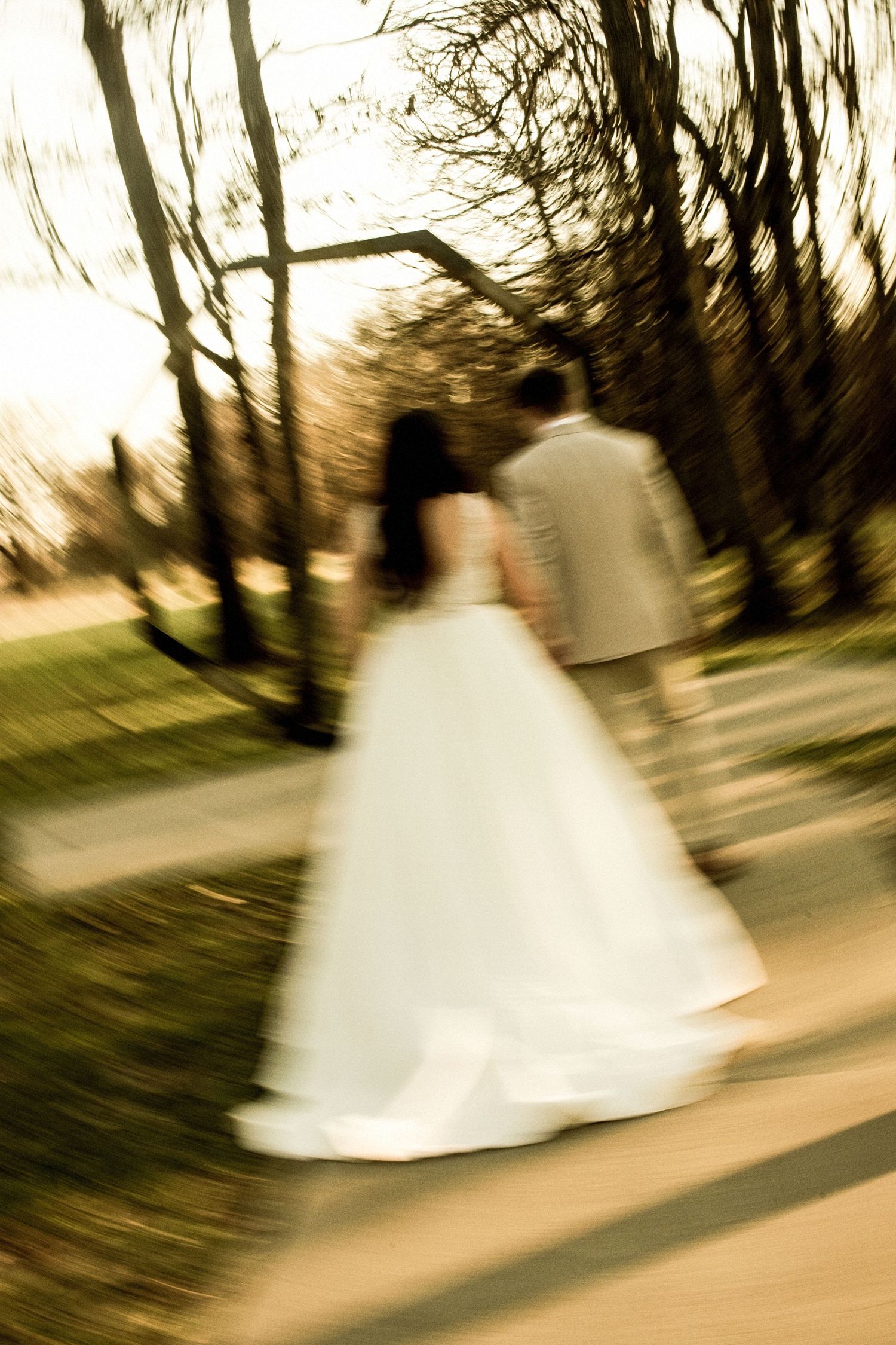 Bride and groom walking hand in hand through tree-lined path, captured with artistic motion blur effect.
