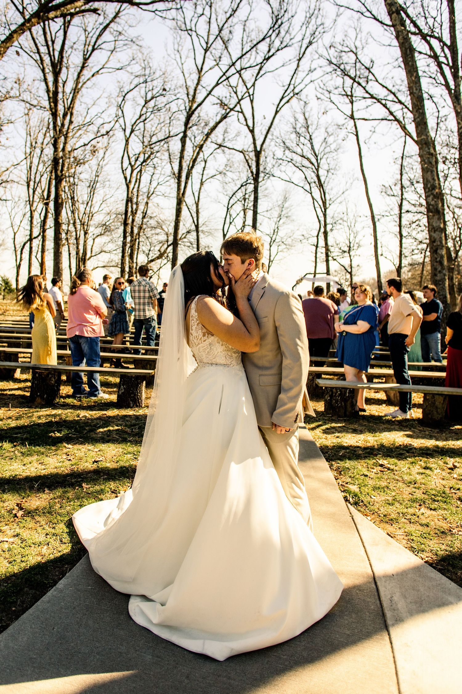 Bride and groom share first kiss at outdoor wedding ceremony surrounded by guests and bare trees.