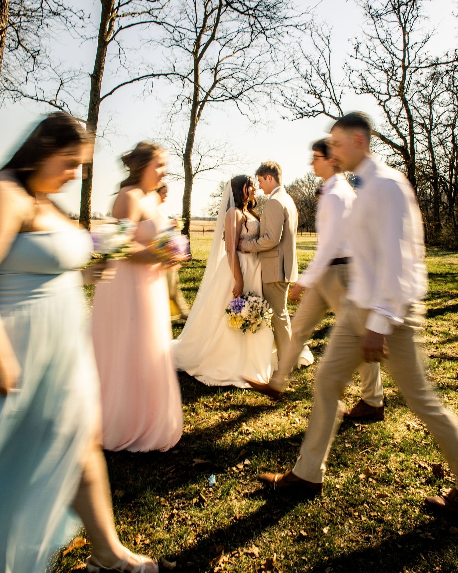 Wedding party walks outdoors among bare trees, bride and groom share a kiss in the center.