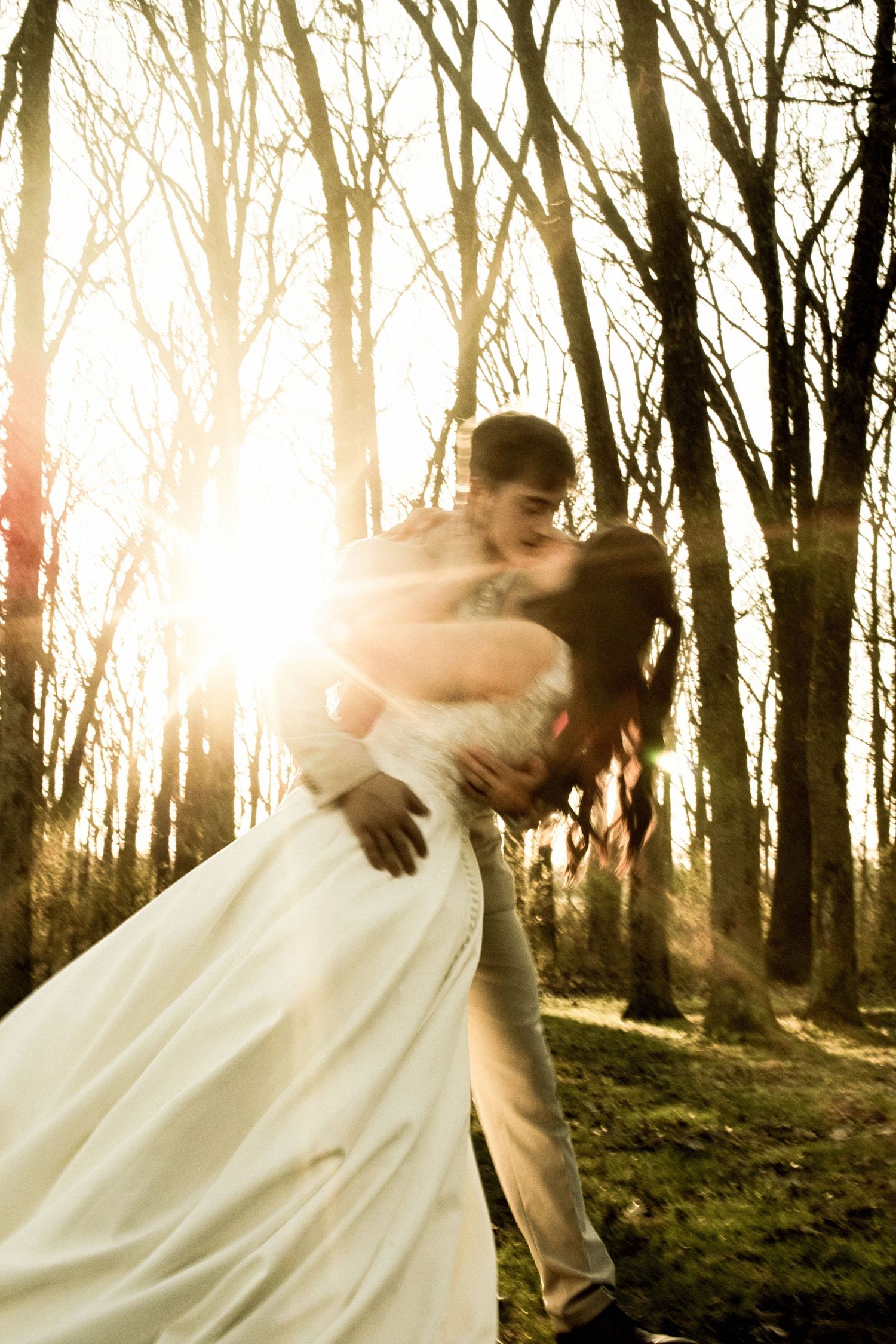 Groom dips bride in white dress for a romantic kiss surrounded by golden sunlight in a forest.