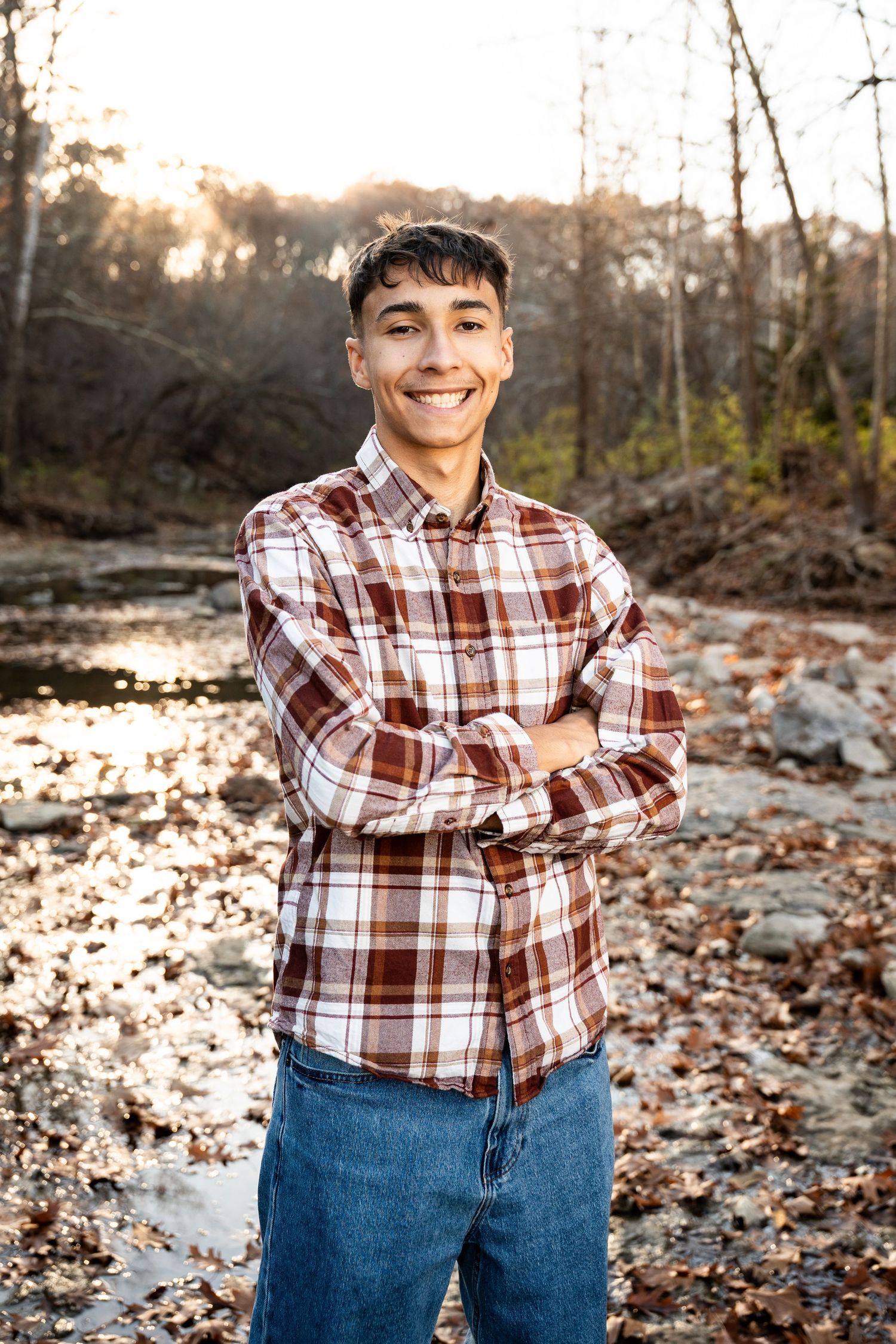 Person in plaid shirt and jeans posing outdoors among fallen autumn leaves.