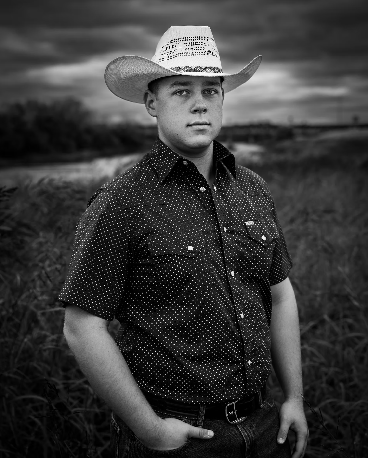 Young person in a polka dot shirt and cowboy hat poses in a dramatic black and white portrait.