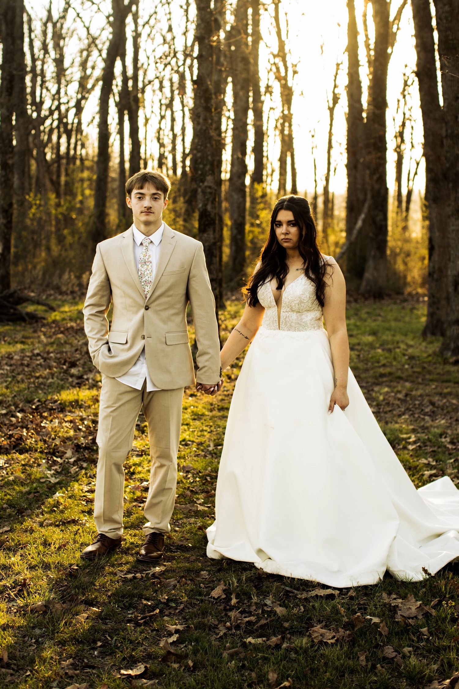 Bride in white gown and groom in tan suit hold hands in sunlit forest during outdoor wedding portrait.