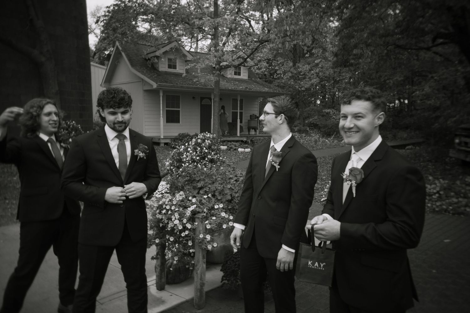 A group of groomsmen in black suits share candid moments while getting ready for a wedding ceremony outdoors.