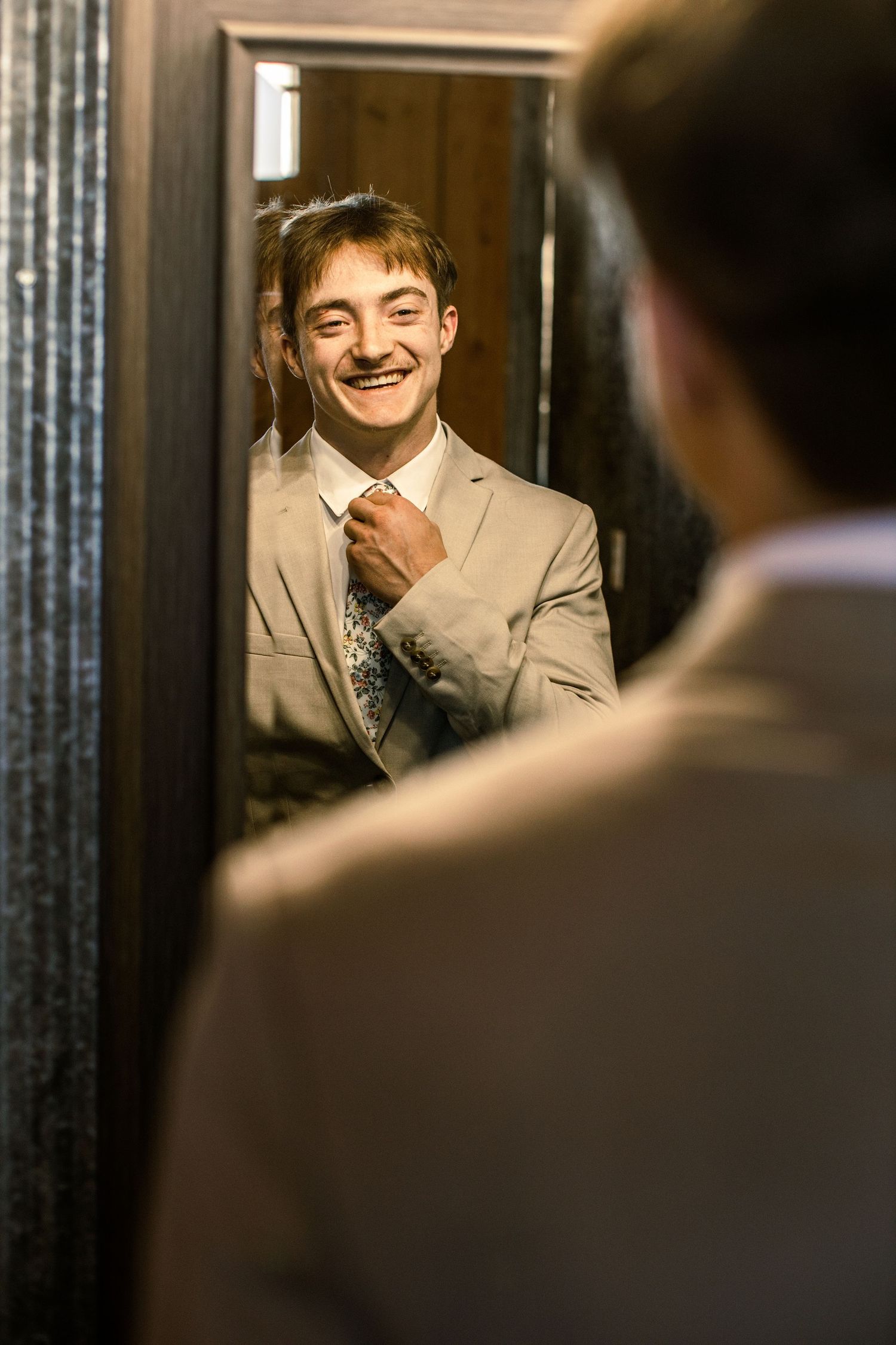 Young groom in tan suit adjusting tie while smiling at his reflection in a mirror before wedding.