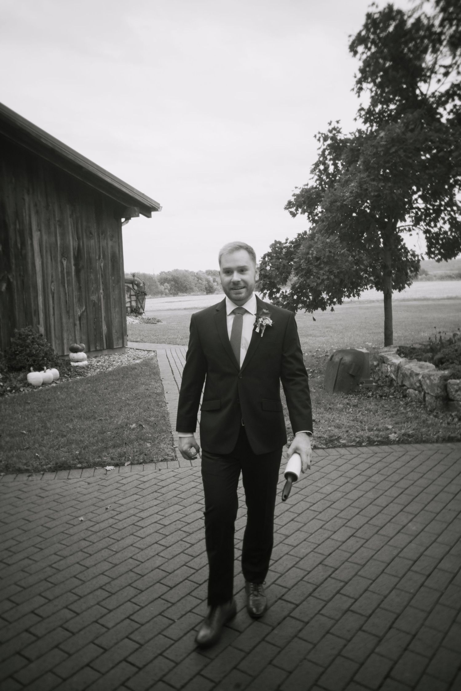 A person in a dark suit walks along a brick paved path beside a rustic barn in a black and white photograph.