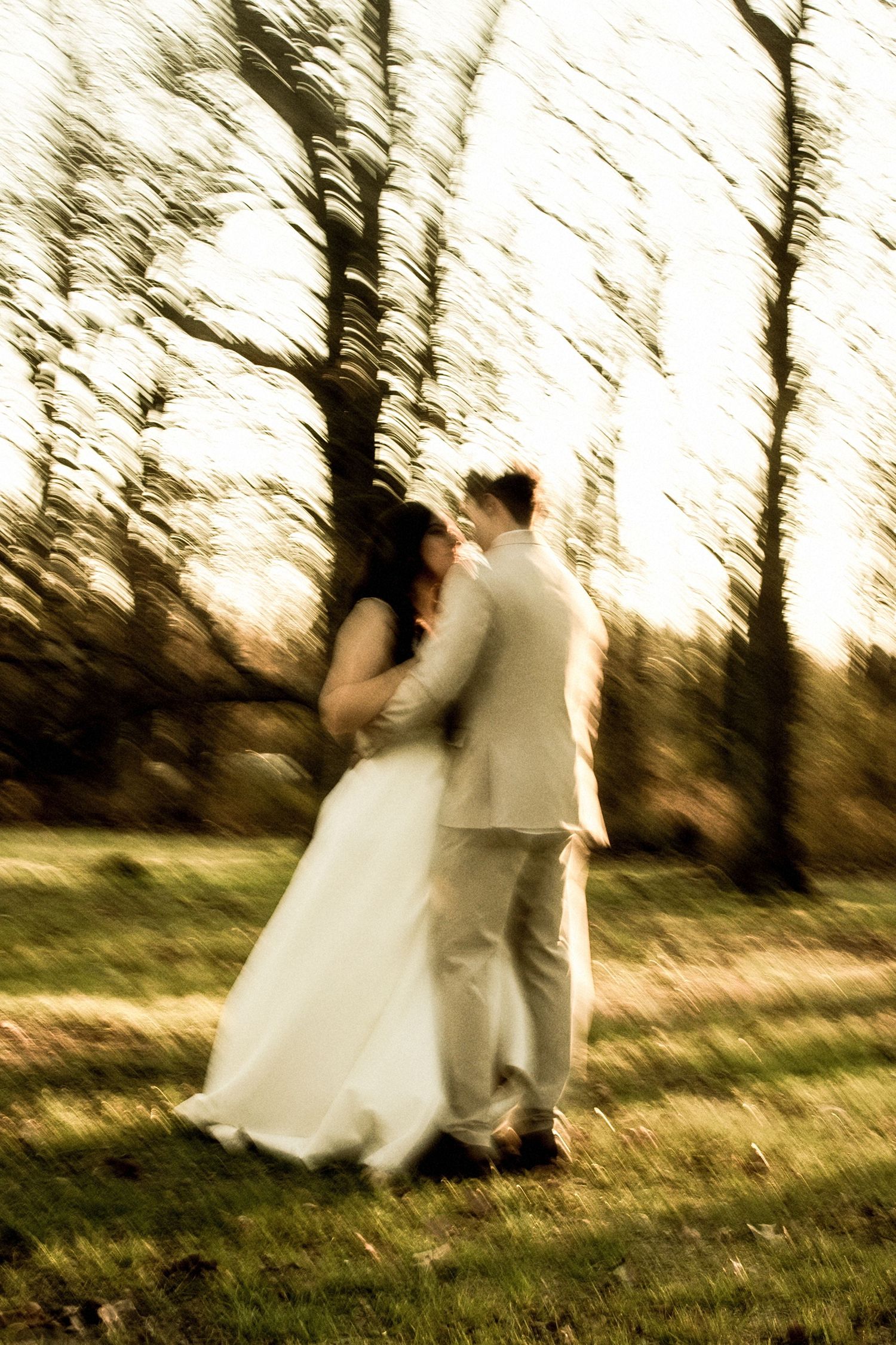 Artistic blurred motion photo of bride and groom embracing in a sunlit outdoor woodland setting.