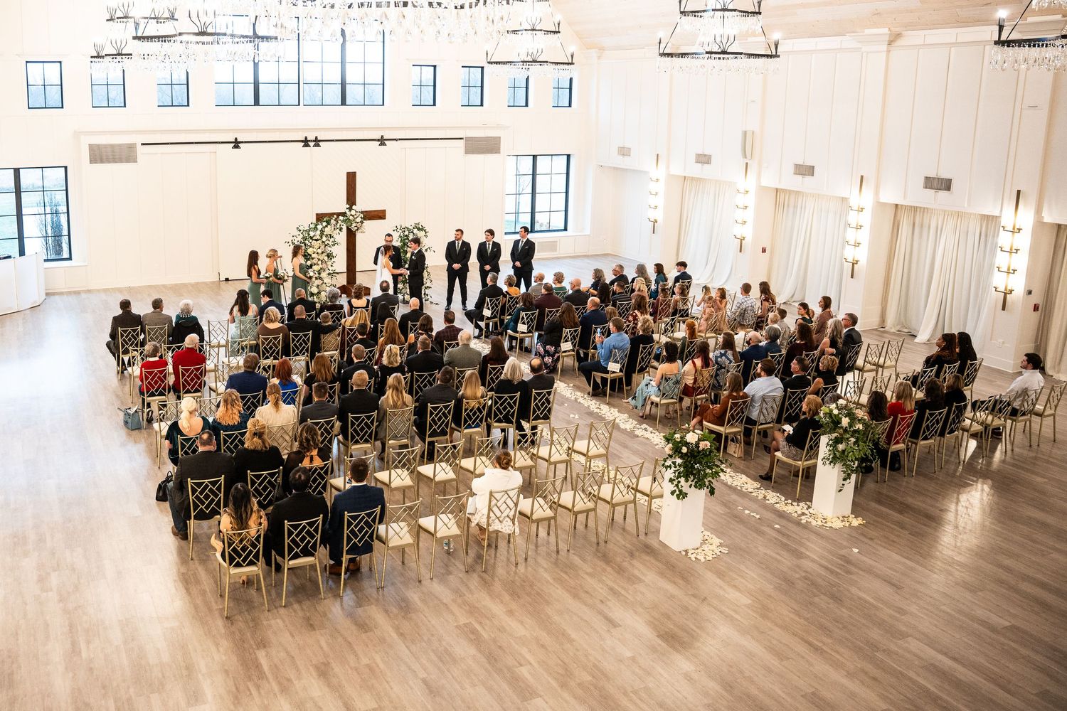 Wedding ceremony in a bright white modern venue with guests seated in rows facing an elegant floral altar setup.