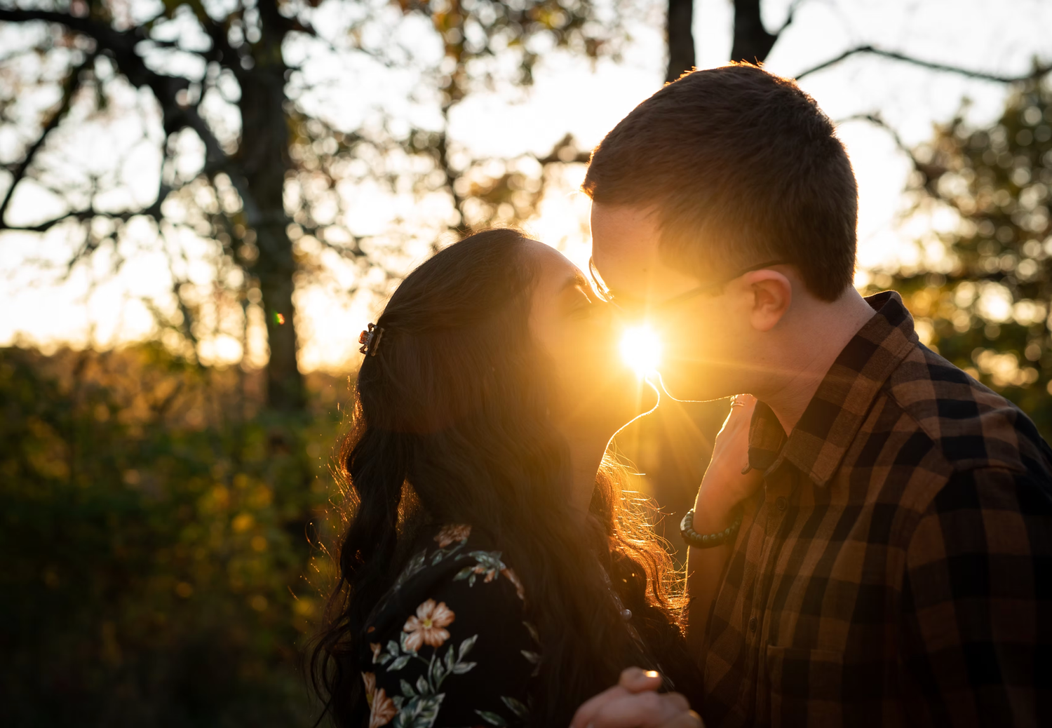 Silhouetted couple embracing during sunset with sun flare through trees.