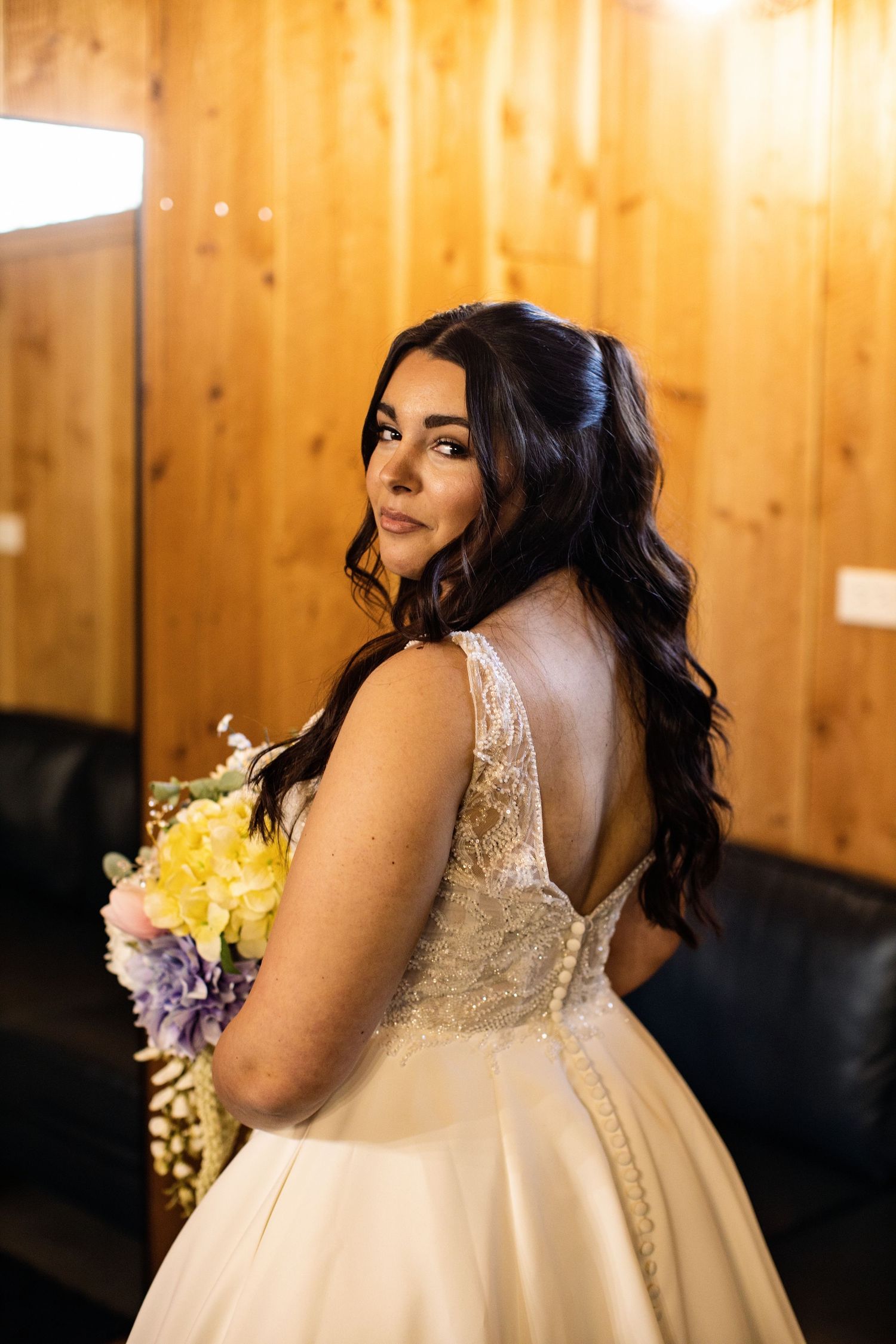 Bride in elegant backless lace wedding gown holding colorful bouquet, posing in rustic wood-paneled room.