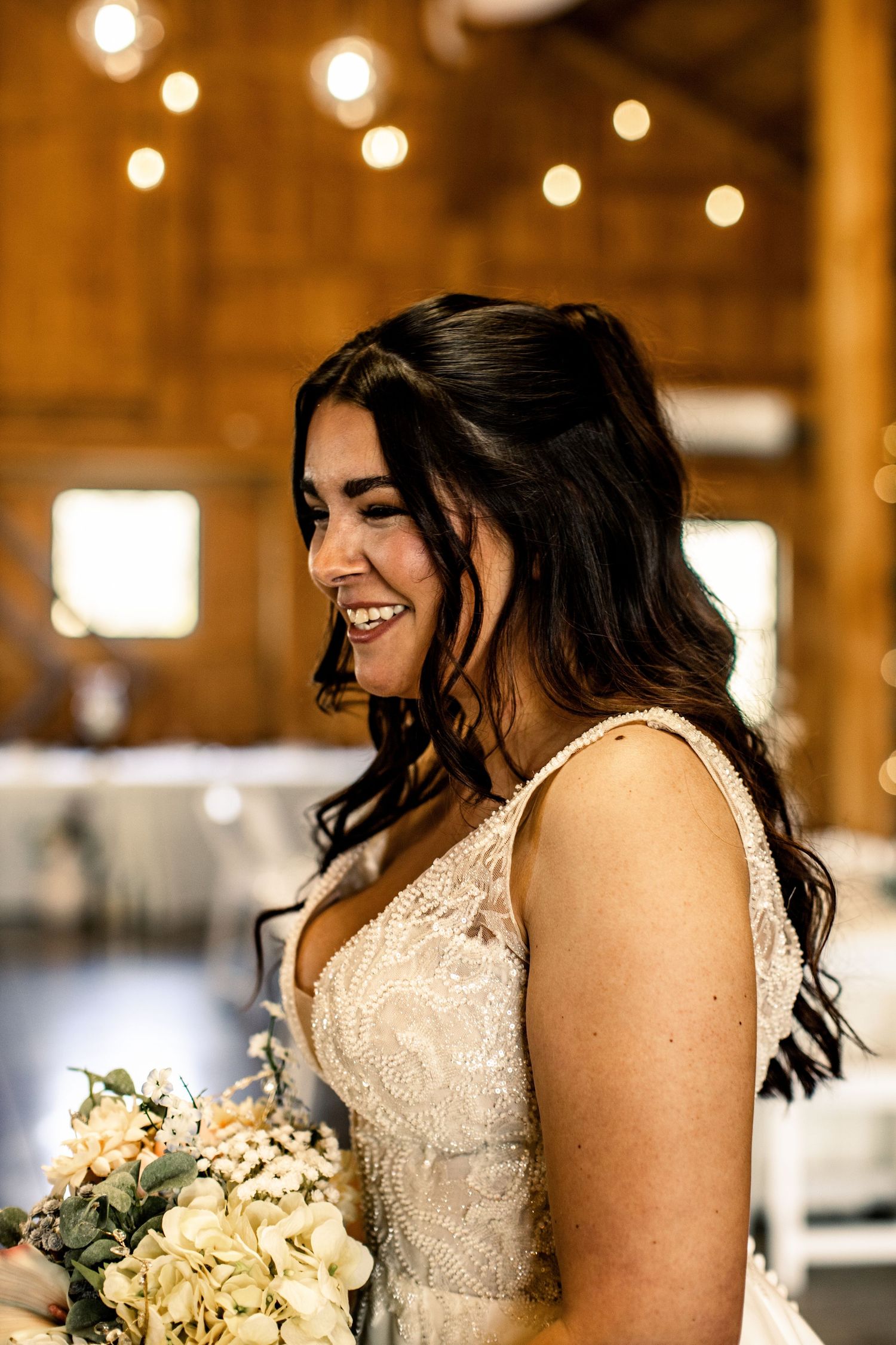 Smiling bride in lace wedding gown holding white floral bouquet inside warmly lit rustic barn venue.