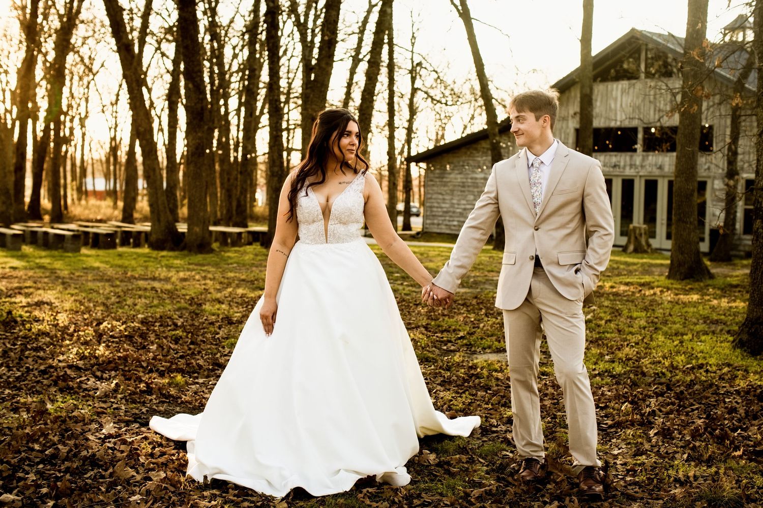 Bride and groom walking hand in hand near a rustic barn in golden sunset light.