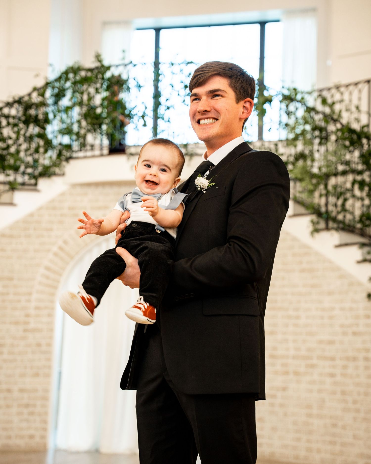 A joyful moment at a wedding venue with someone in a black tuxedo holding a laughing baby in formal attire.
