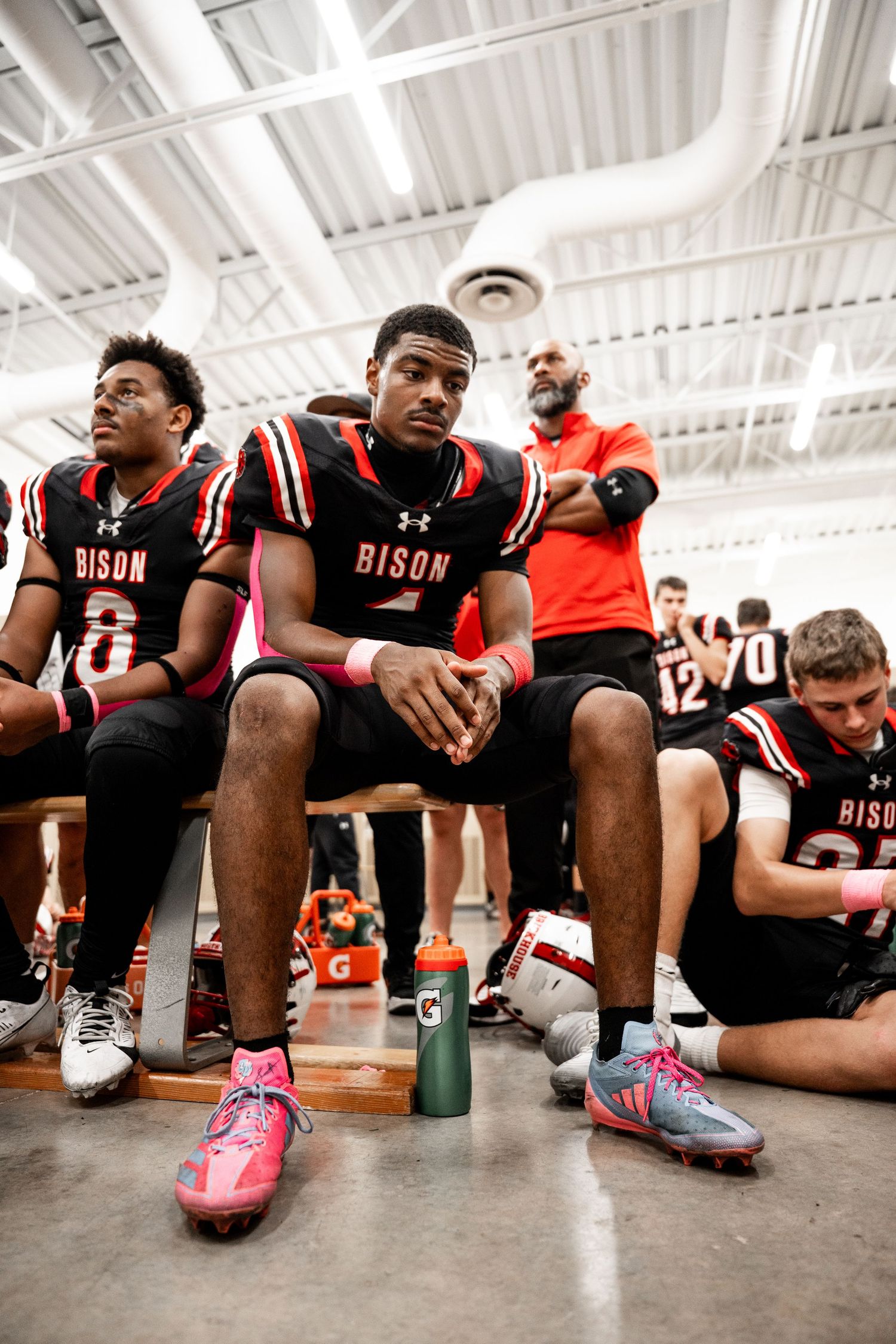 Football players in black Bison uniforms sitting on bench showing their cleats with pink accents.
