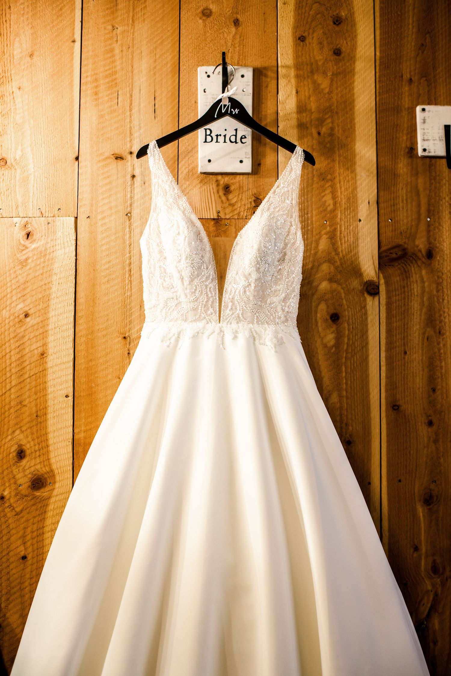 White lace wedding gown hanging on a bride hanger against a rustic wood wall.