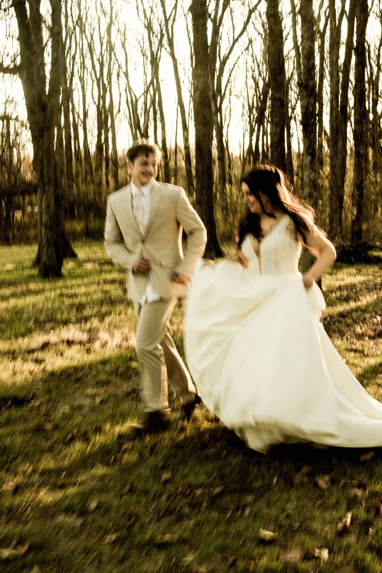 Bride and groom playfully running through sunlit forest in wedding attire on their special day.