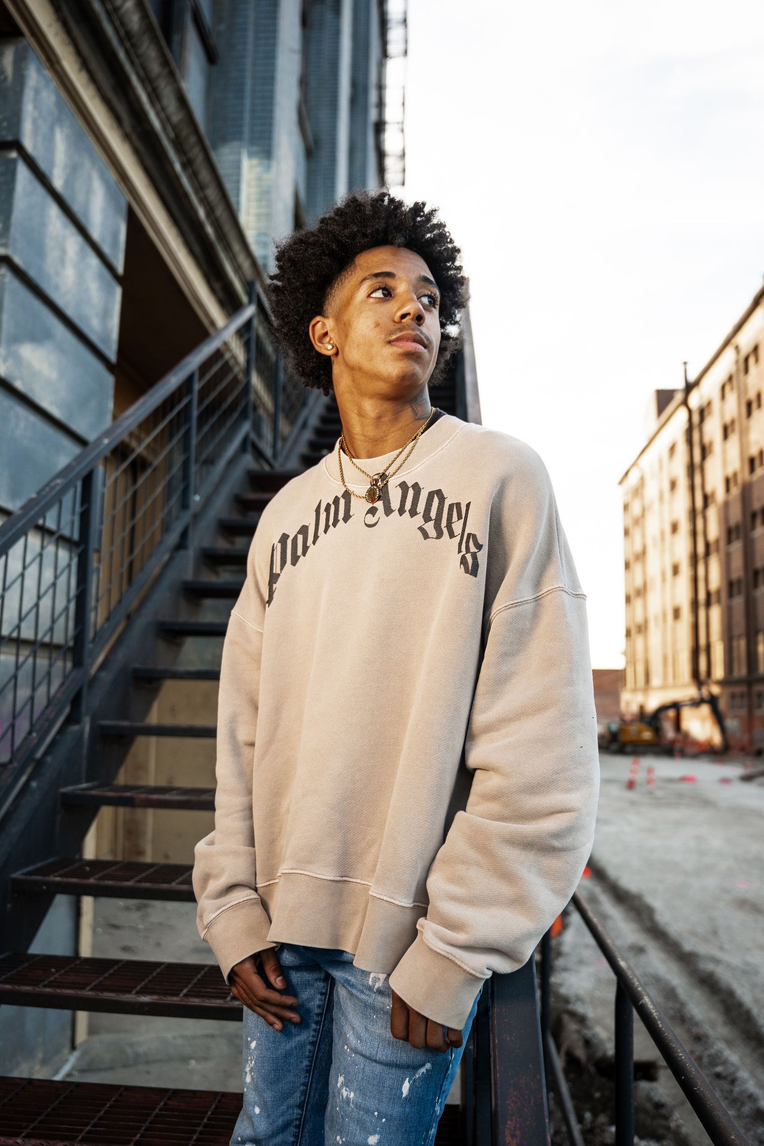 A person in a white sweatshirt and jeans poses against an urban backdrop with fire escapes and buildings.