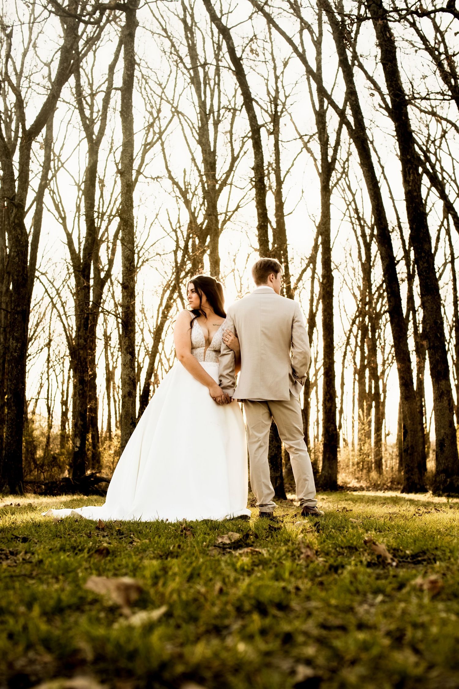 Bride and groom hold hands while standing among bare trees in a sun-drenched forest wedding photo.