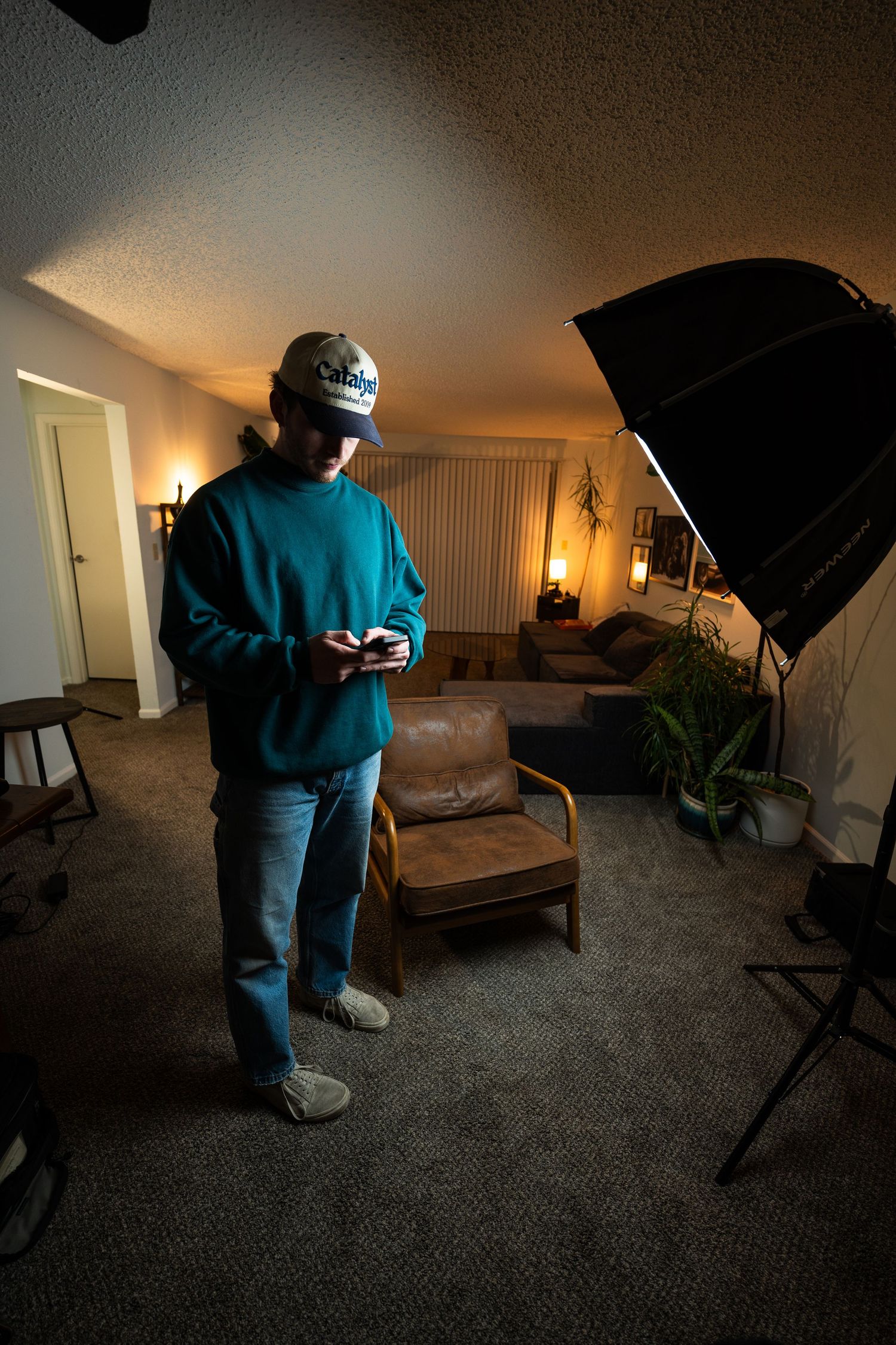 A photographer in blue clothing adjusts lighting equipment in a dimly lit room with a couch and studio setup.