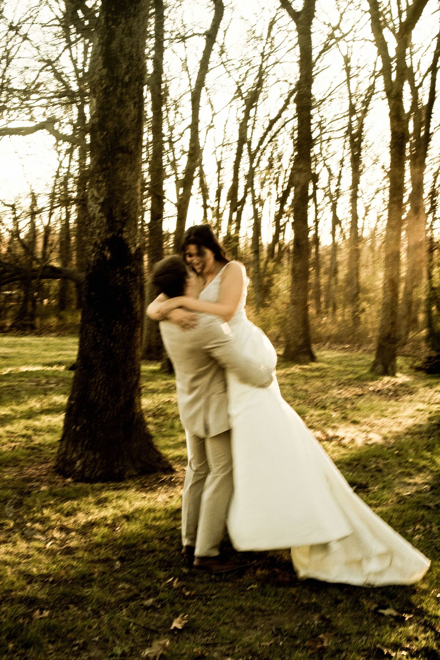 Bride and groom embracing and laughing in golden sunset light among bare trees during wedding portraits.