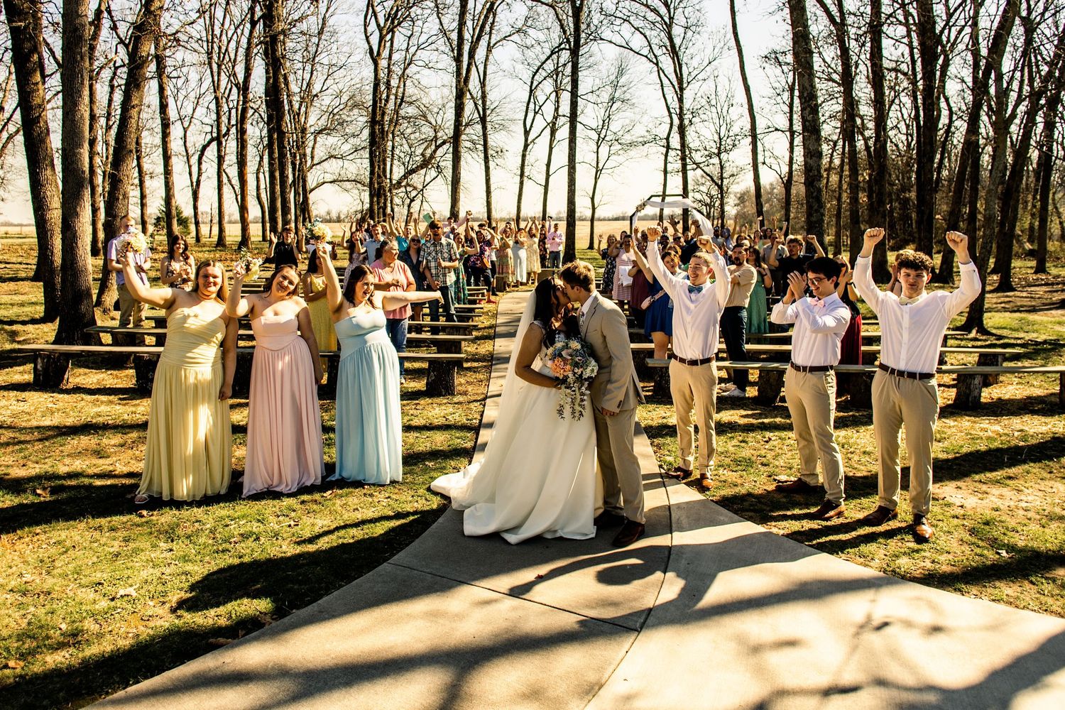 Outdoor wedding ceremony with bride and groom kissing, surrounded by cheering wedding party in colorful dresses and suits.