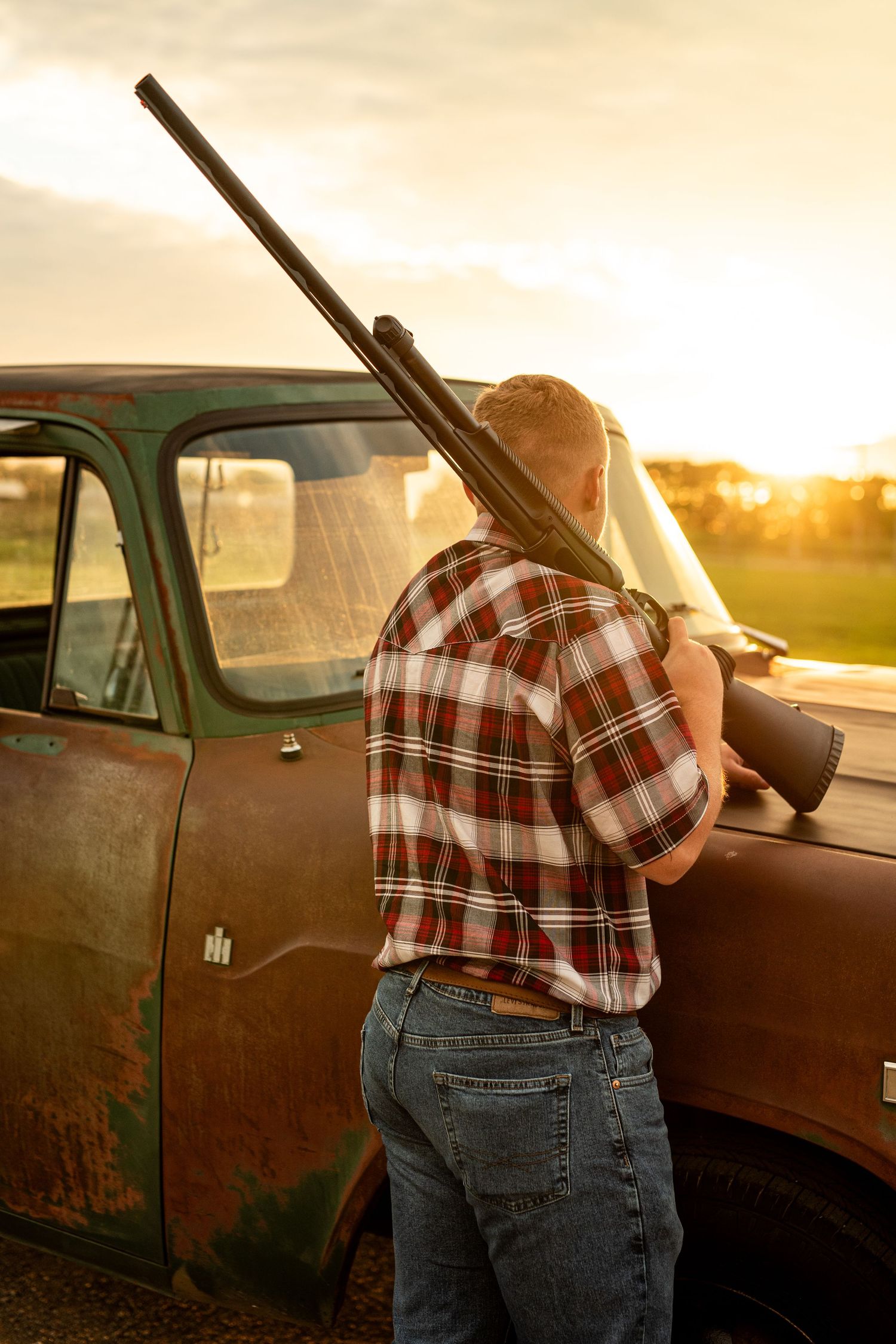 Person in plaid shirt leaning against rustic brown truck at sunset.