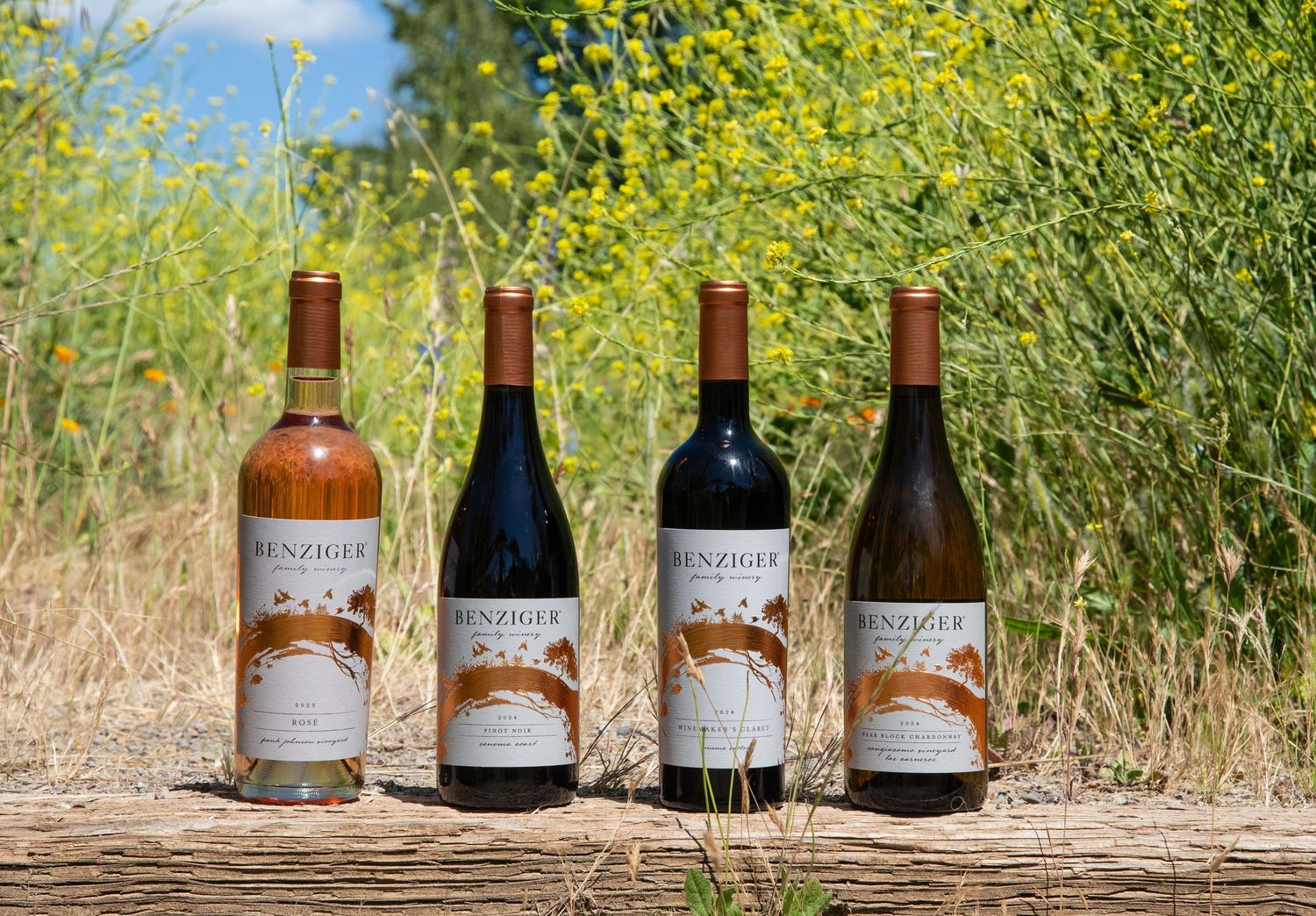 Four Benziger wine bottles displayed on a wooden surface with vibrant yellow wildflowers and blue sky in the background.