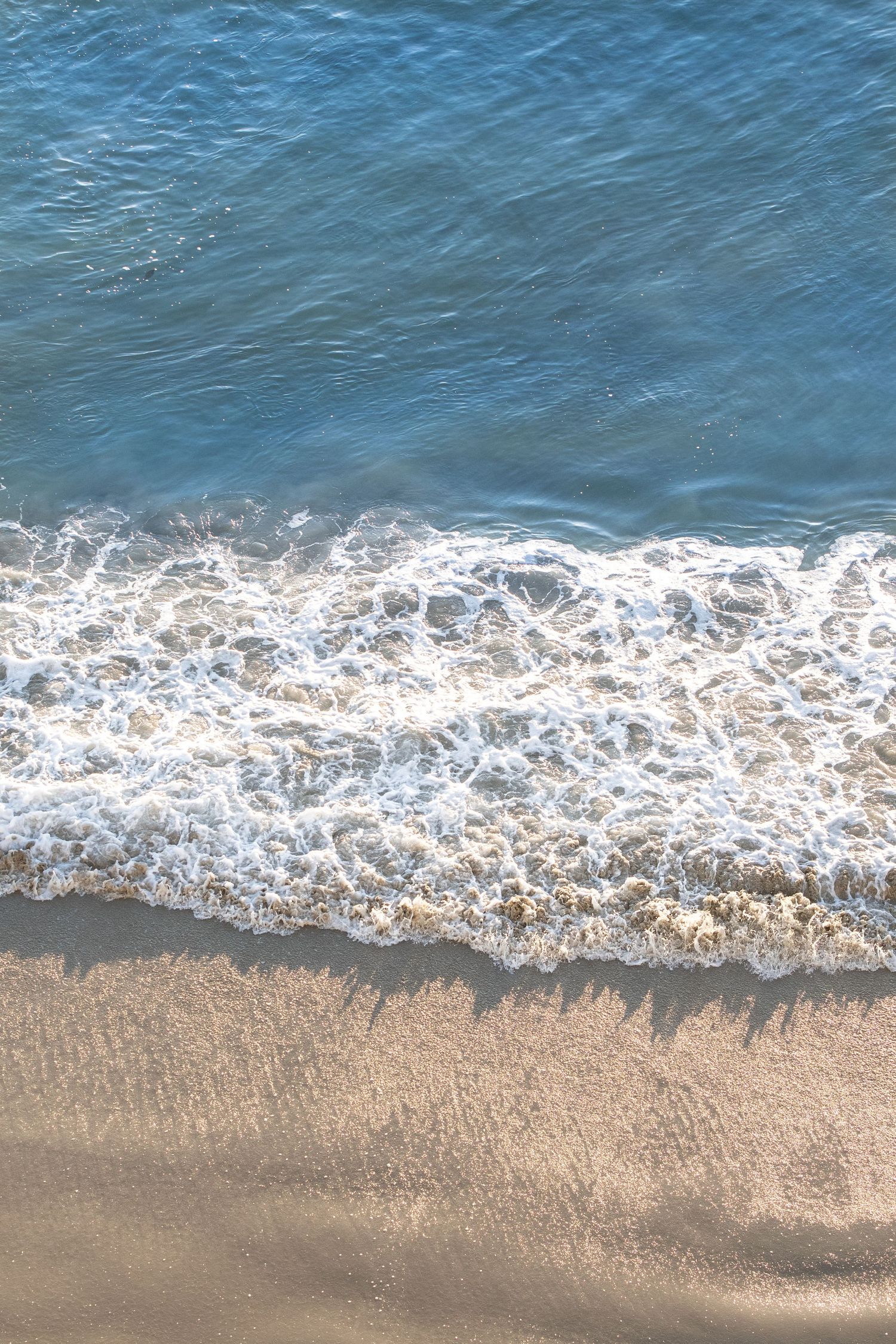 Aerial view of ocean waves meeting sandy beach with gentle foam patterns.