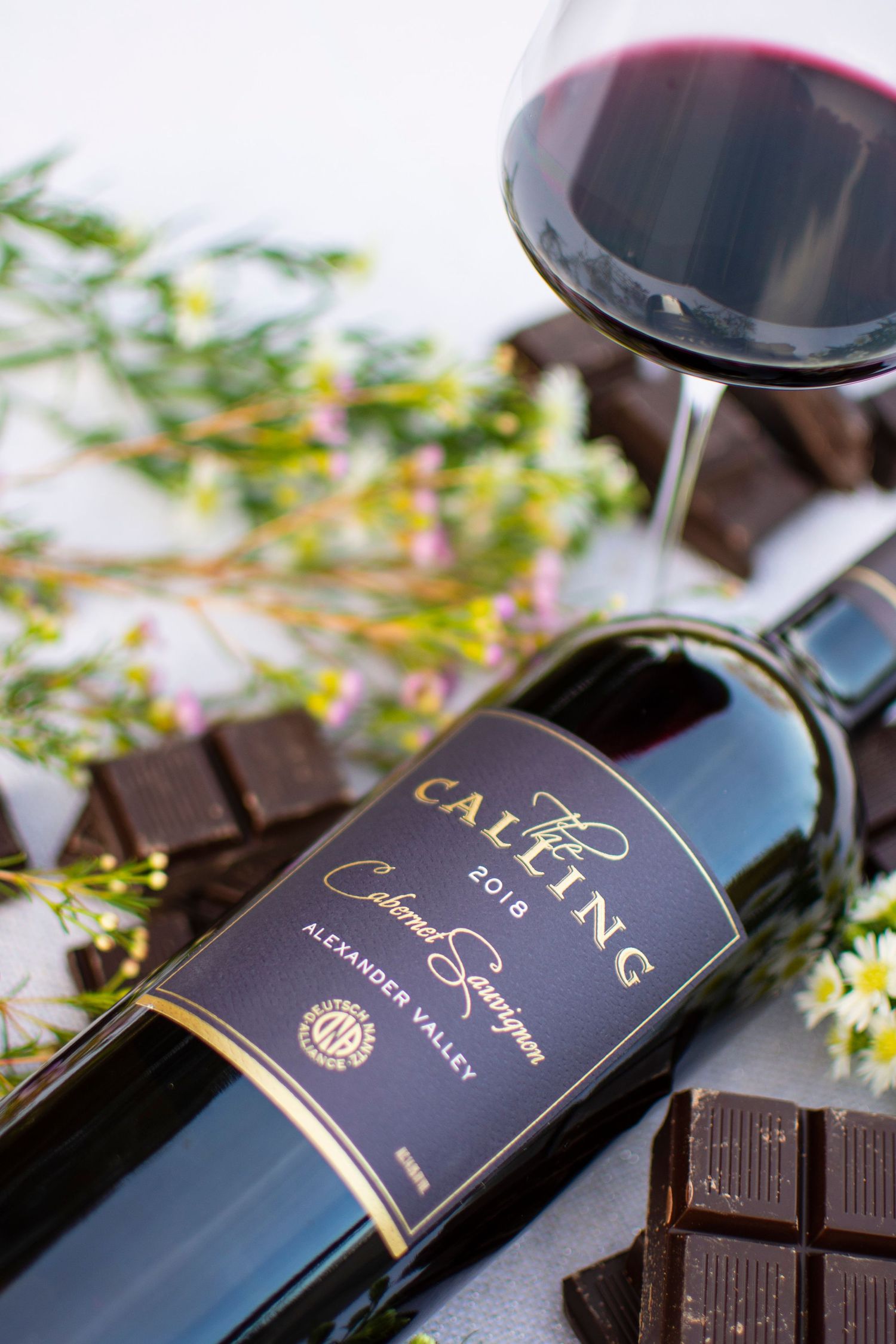 Bottle of Caymus wine with a glass of red wine and dark chocolate on a decorated surface.