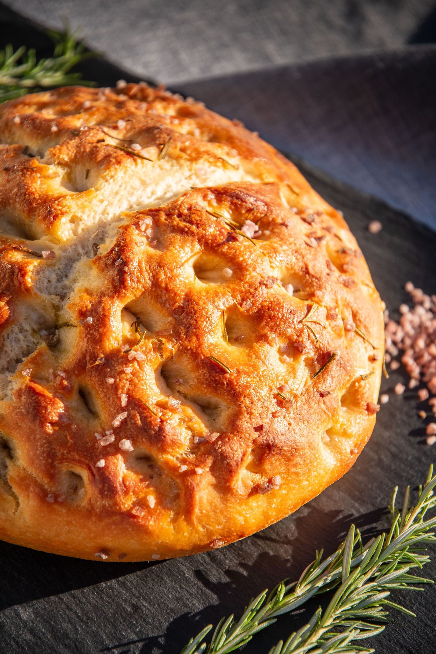 Golden brown artisanal bread with dimpled surface and rosemary garnish on slate board.