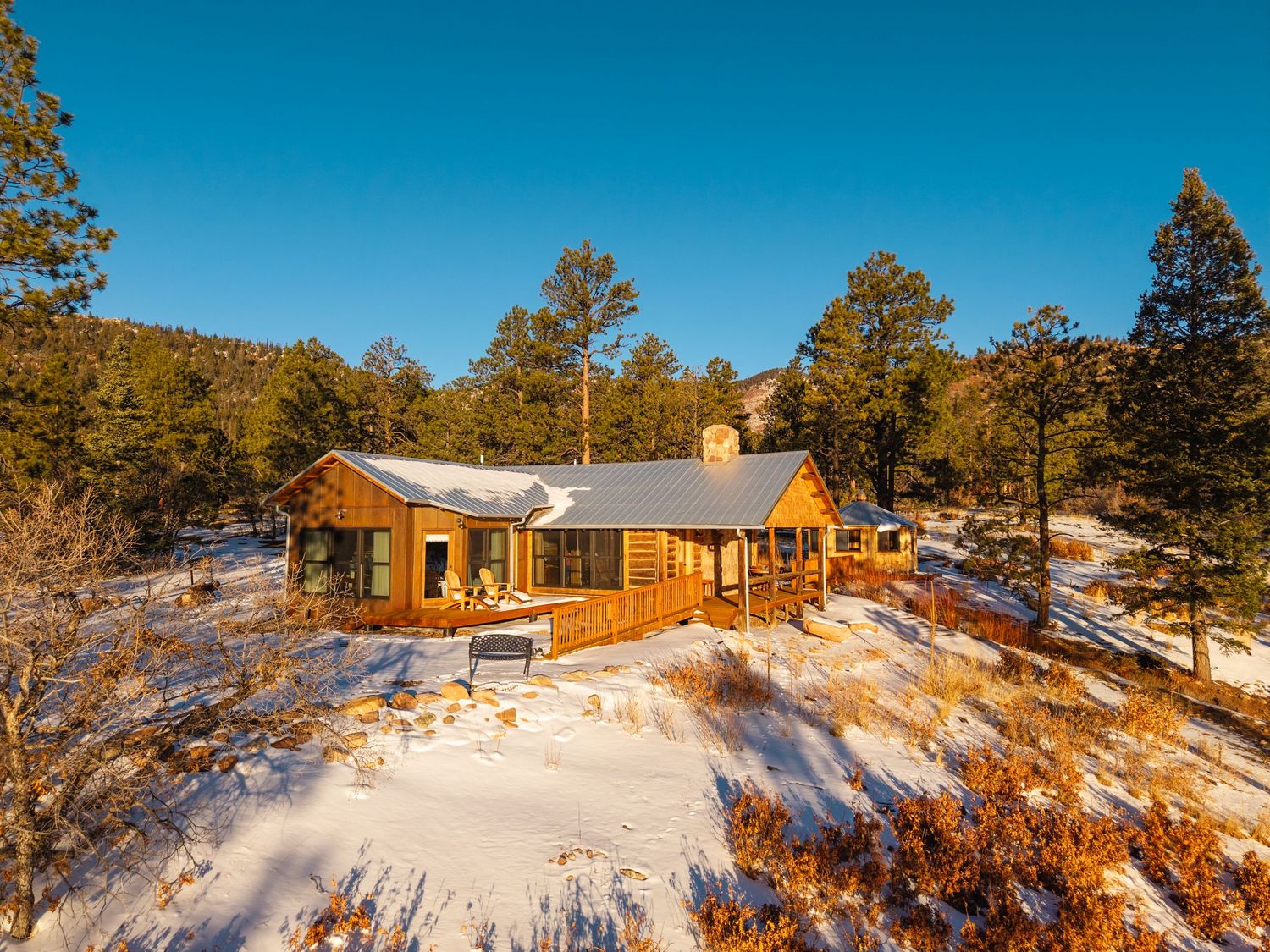 Rustic log cabin nestled among pine trees in a snowy winter landscape under a clear blue sky.