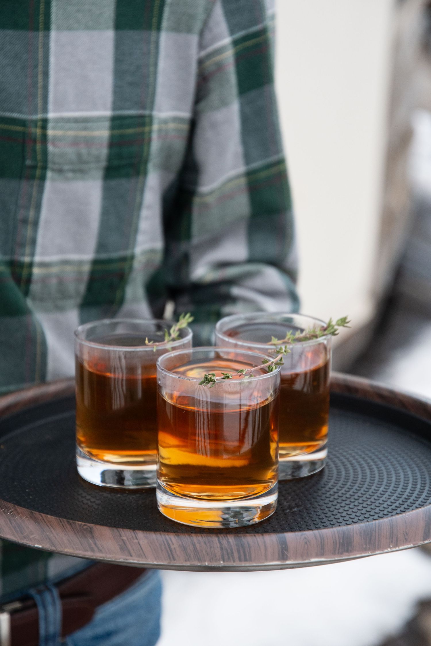 Three amber-colored cocktails with herb garnishes served on a dark serving tray.