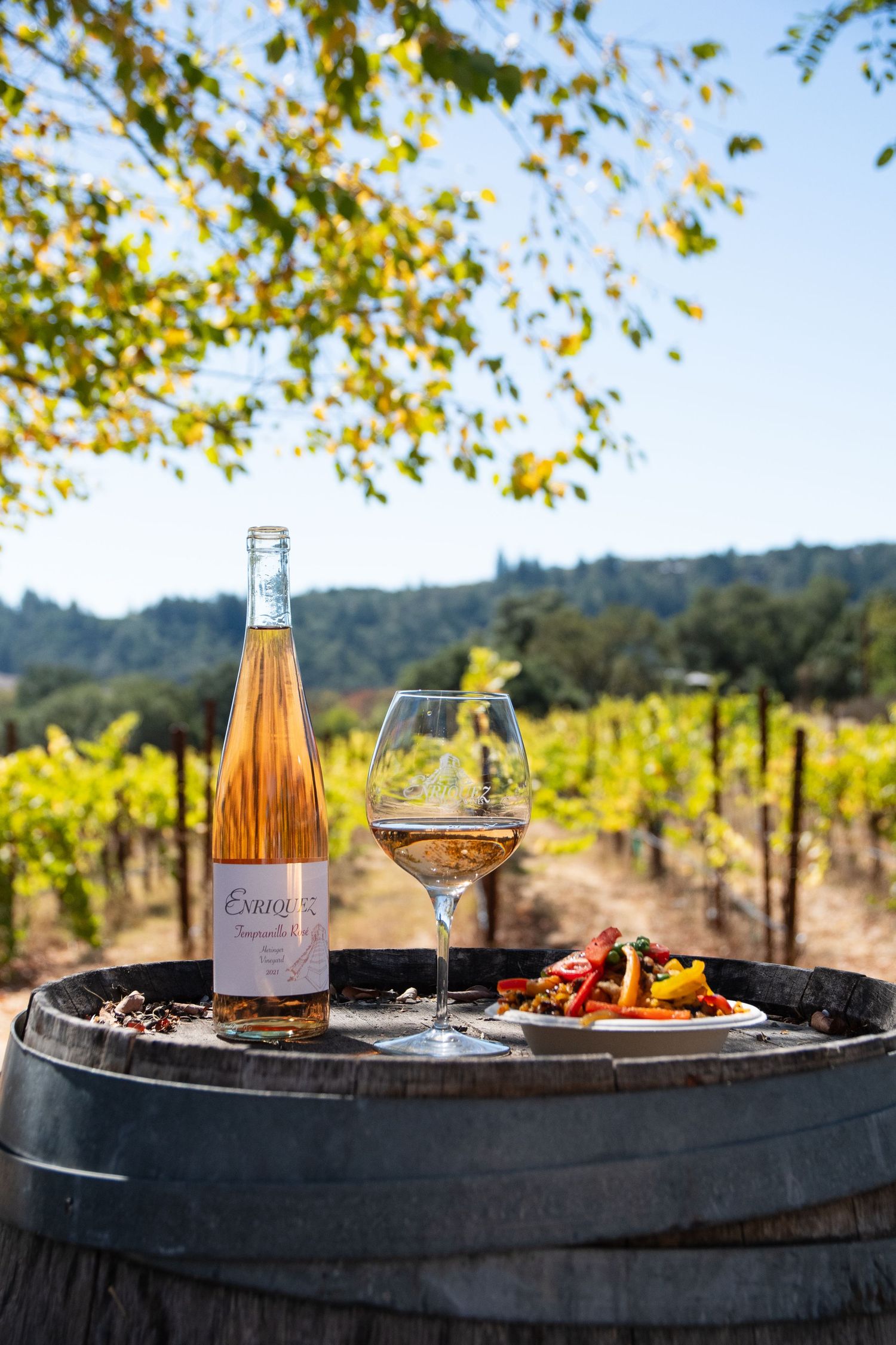 Rose wine bottle and glass on a wooden barrel amid lush vineyard rows, paired with a colorful roasted vegetable dish.