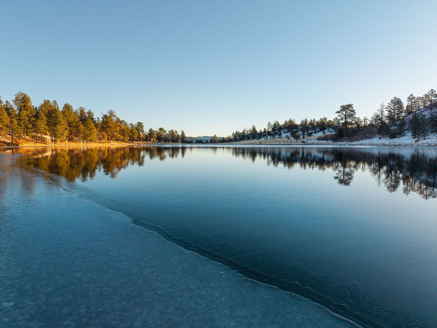 Calm mountain lake with partially frozen icy edge reflecting pine trees and a clear blue sky at sunrise.