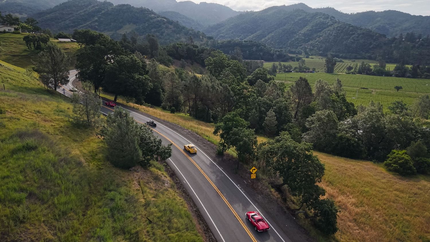 Aerial view of cars driving on a winding road through lush green hills and mountains in a scenic rural landscape.
