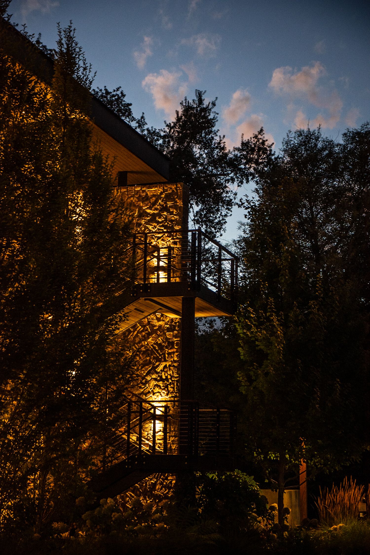 Warm light glows through windows of a modern wooden house nestled among trees at dusk.