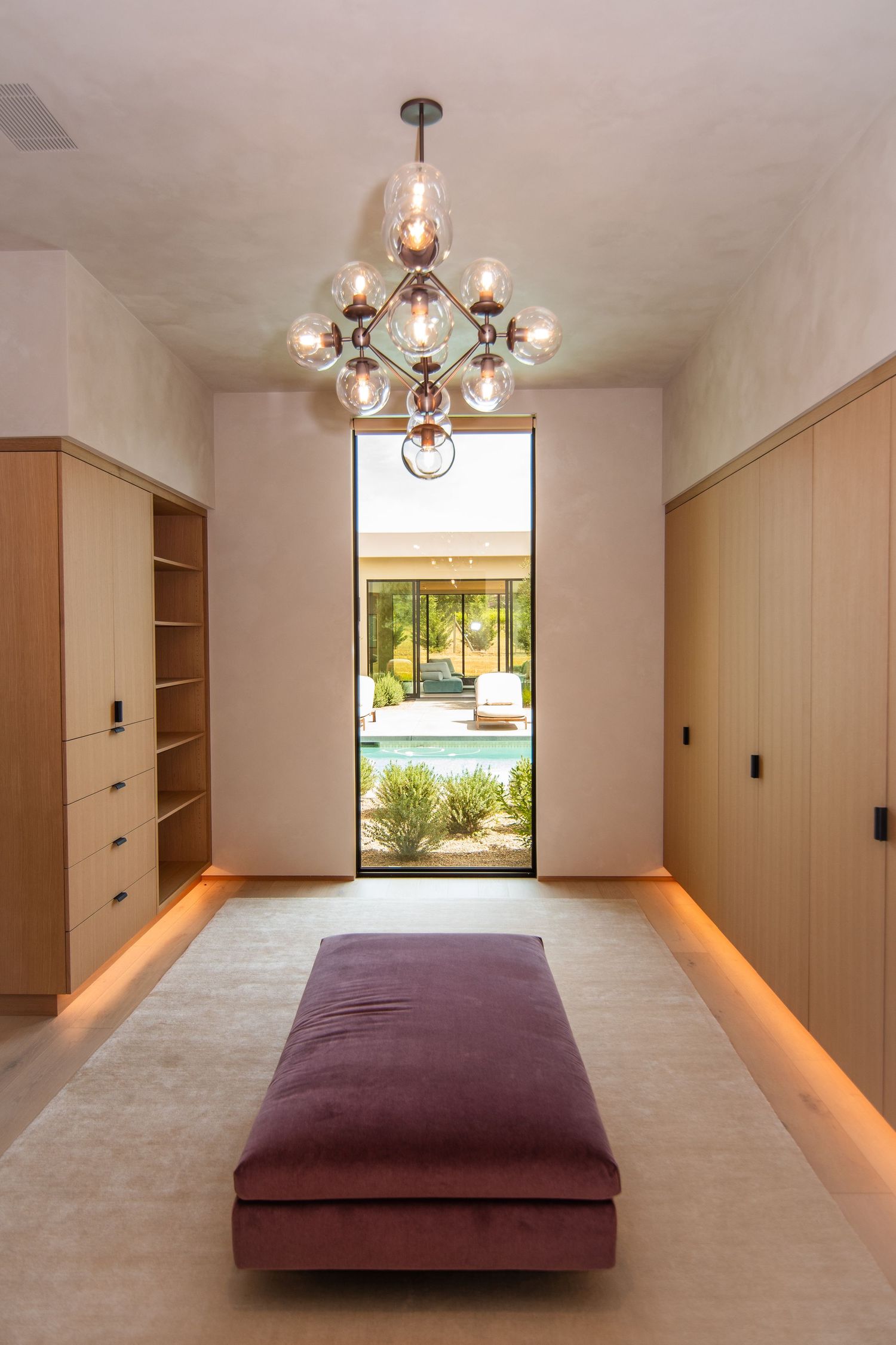 Modern walk-in closet with plush bench and modern chandelier featuring a view of the pool.