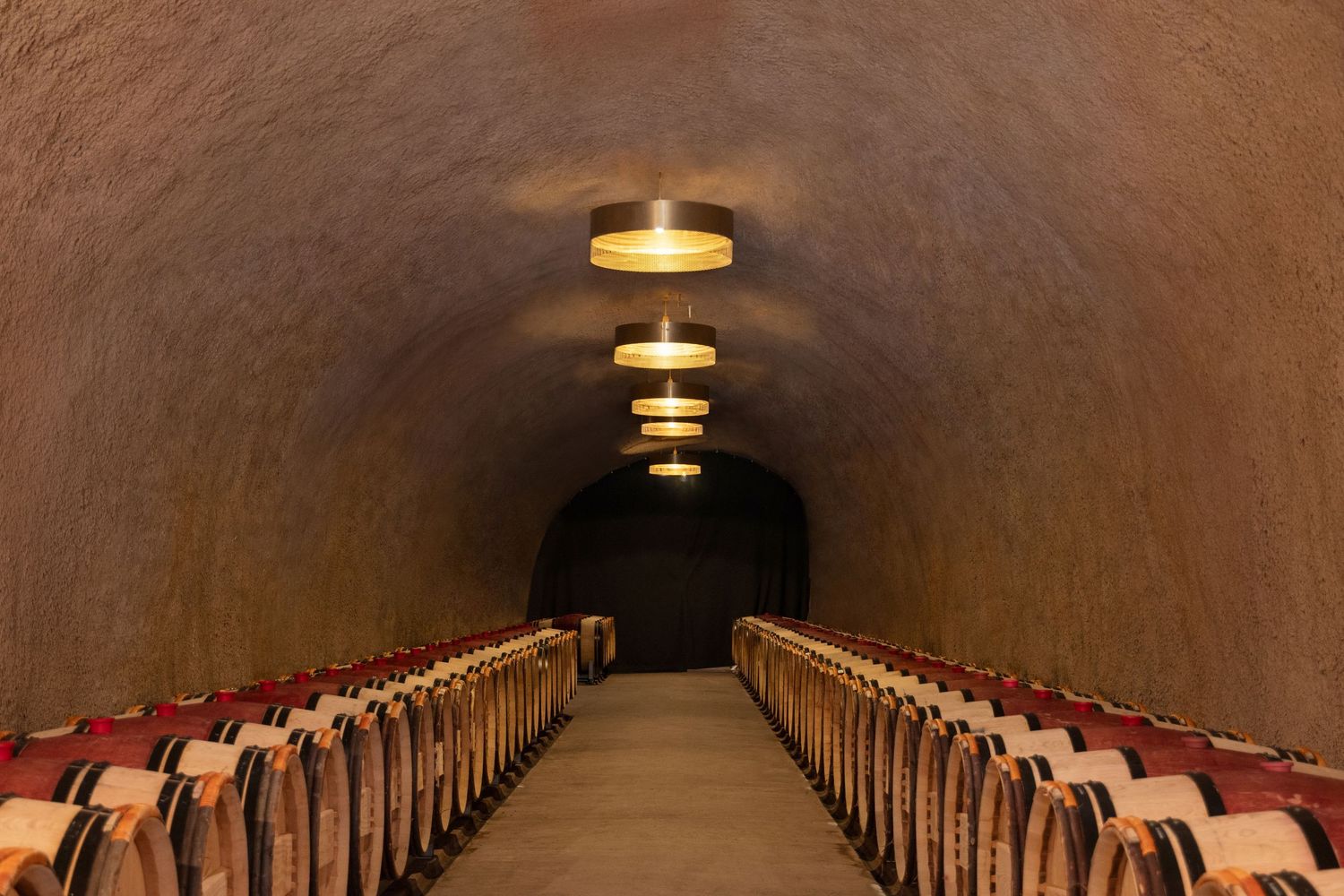 A symmetrical wine cellar tunnel with rows of oak barrels and elegant ceiling lights receding into the distance.