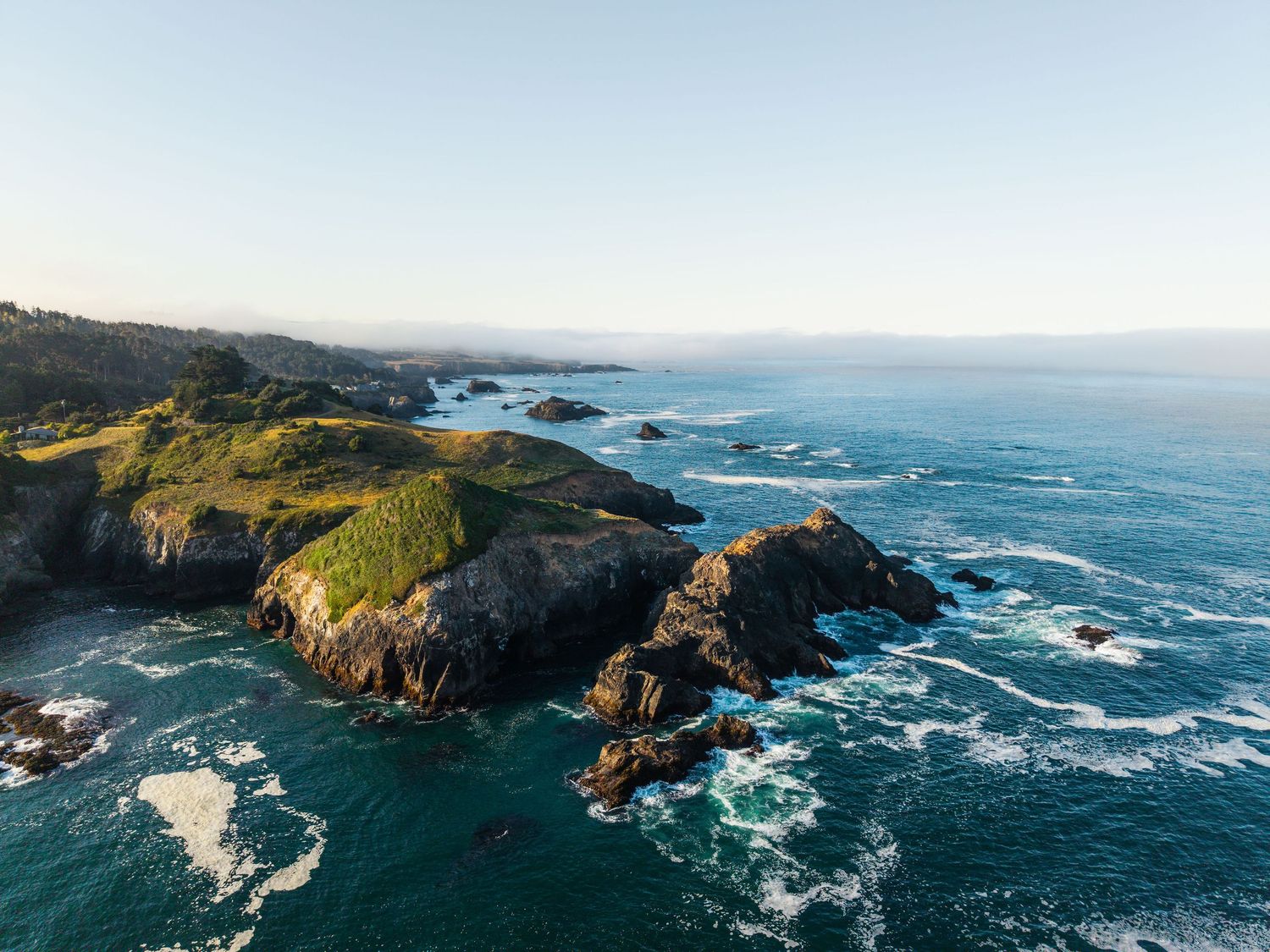 Aerial view of rocky coastal cliffs with lush green hills meeting turquoise ocean waves under a clear blue sky.