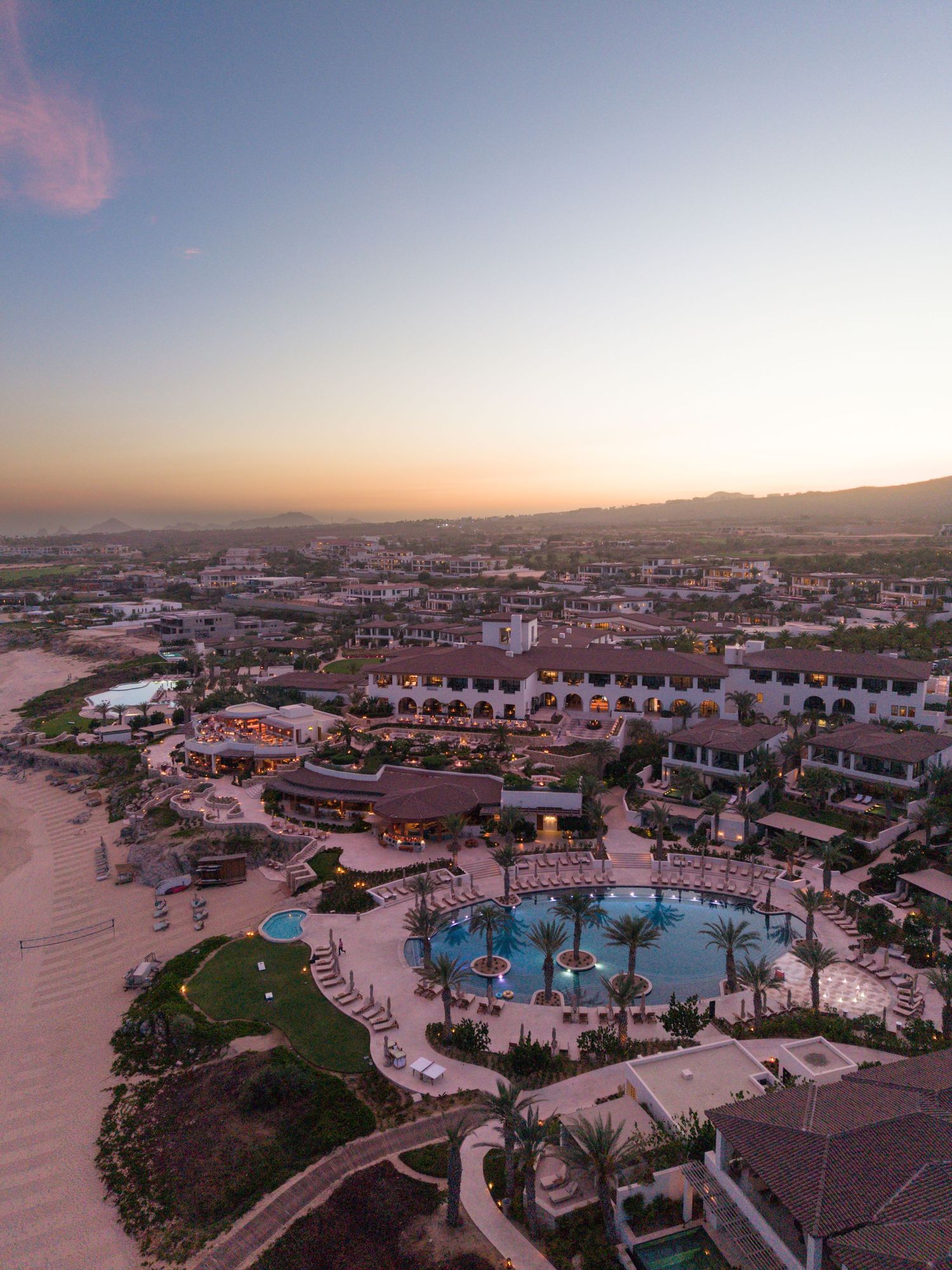 Aerial sunset view of a luxury resort with pools, palm trees, and sandy beach in a coastal town.