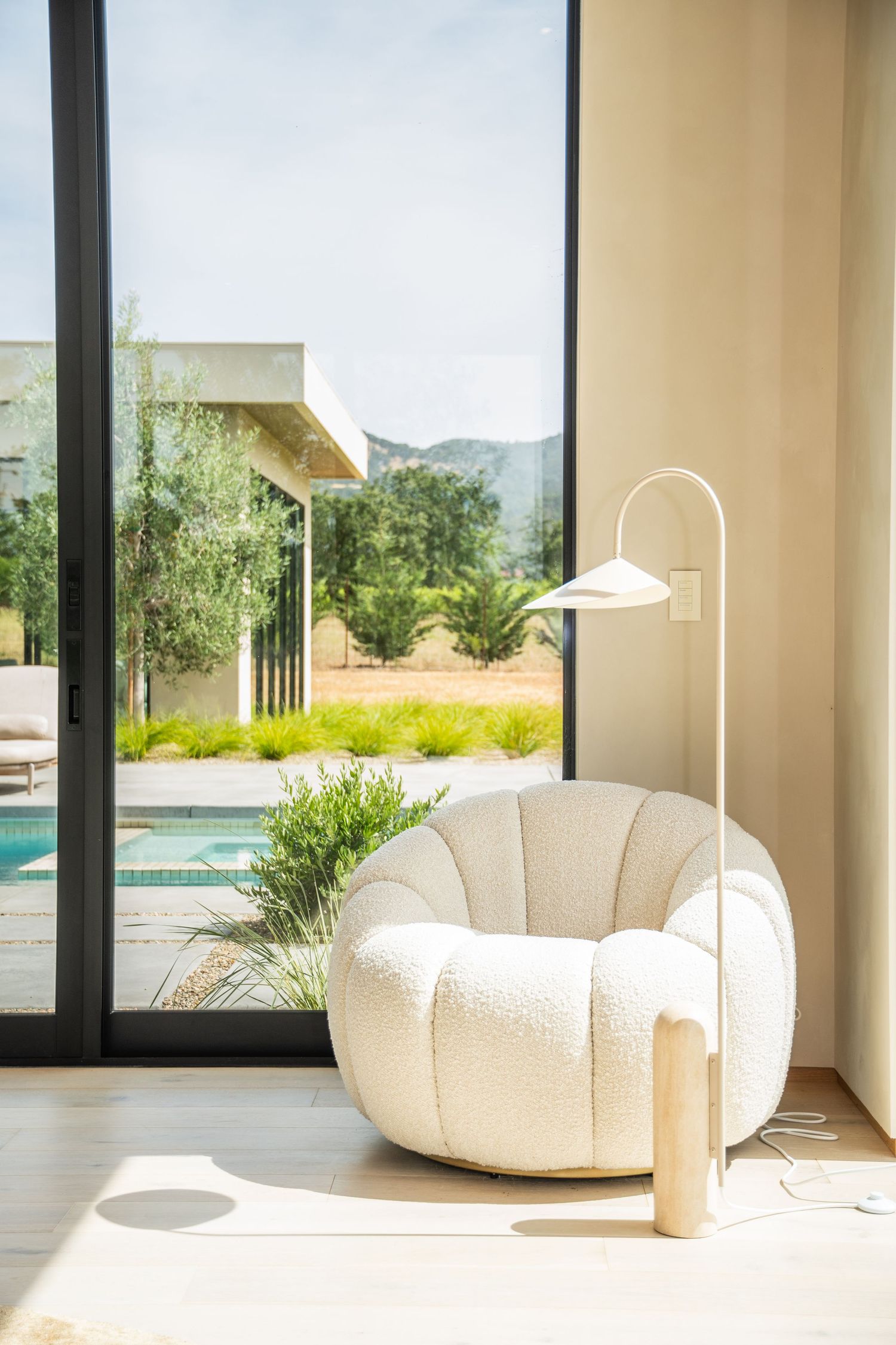 Modern cream-colored accent chair with arched lamp beside floor-to-ceiling windows overlooking pool.