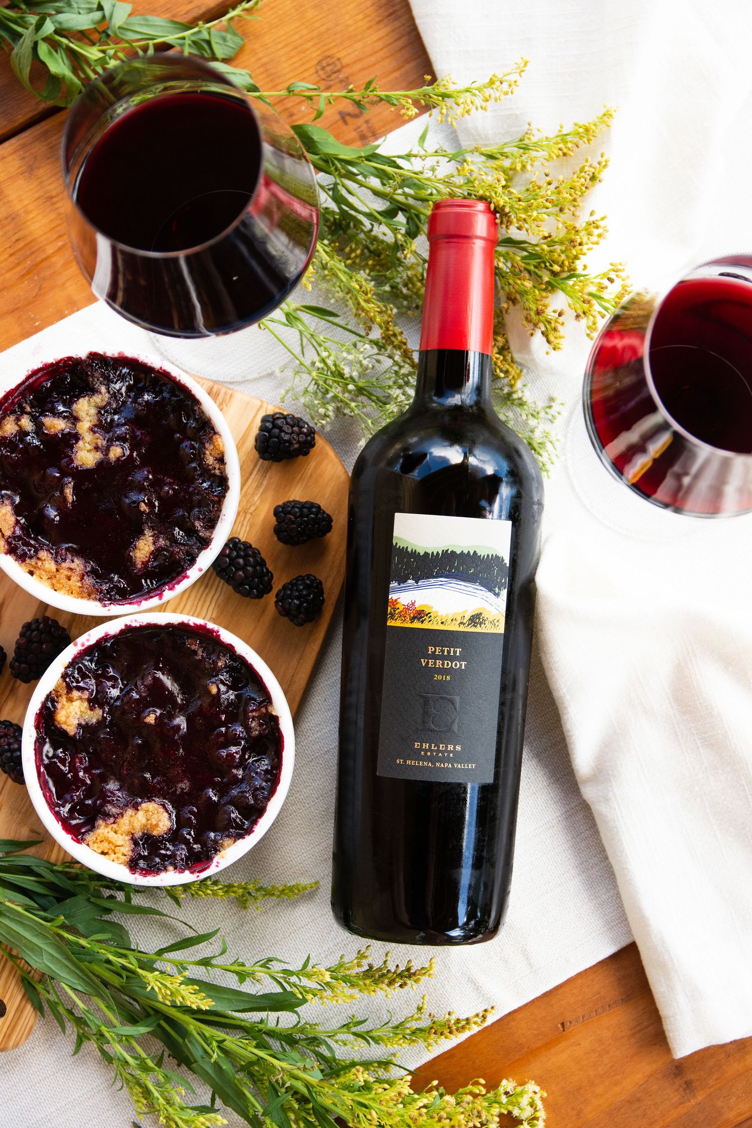 Flatlay of wine bottles and blackberry desserts on wooden surface with fresh herbs.