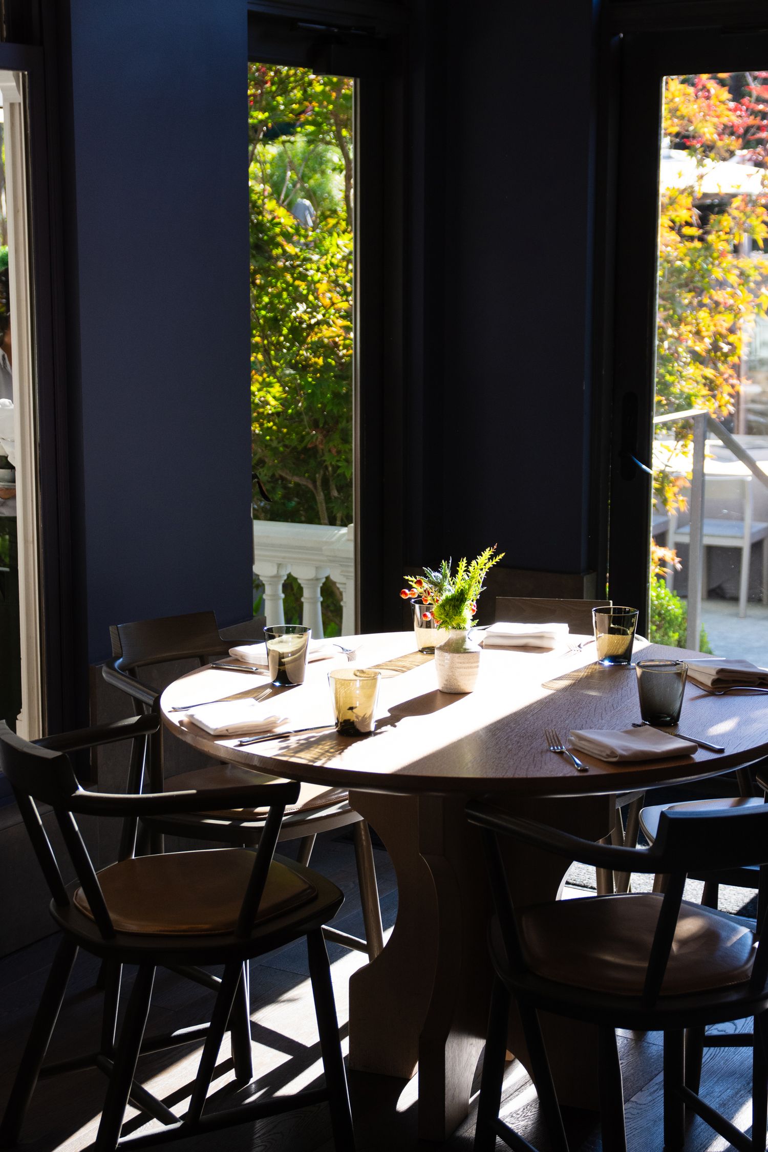 Restaurant interior with sunlit tables by windows overlooking autumn foliage.