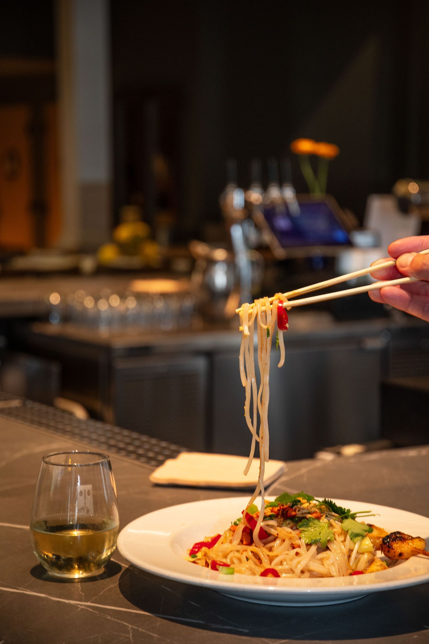 Chopsticks lifting noodles from an elegantly plated pasta dish with a glass of white wine on a dark restaurant table.