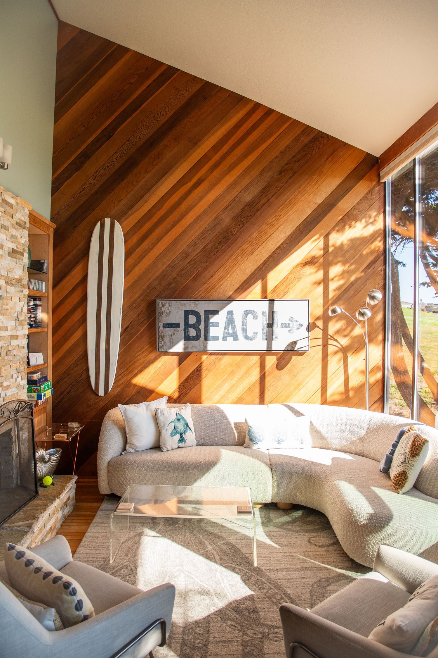 Bright beach-themed living room with wood-paneled walls, surfboard decor, curved white sofa, and a BEACH sign.
