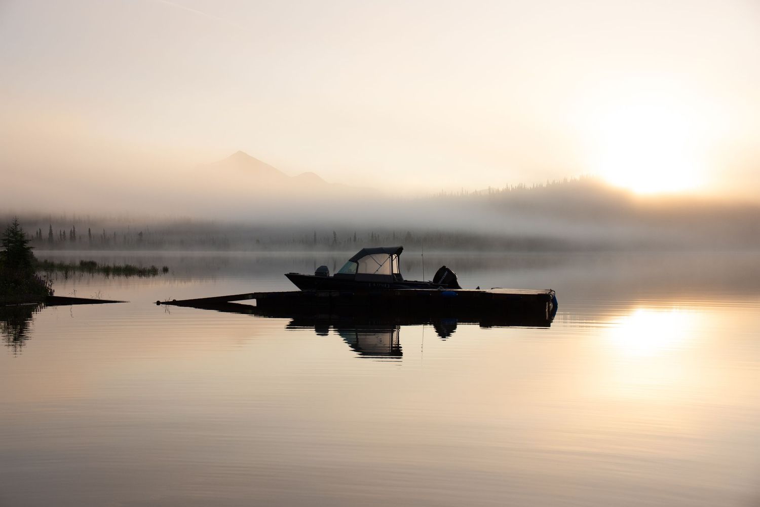 Silhouette of a boat on a misty lake at sunrise with fog-covered mountains and golden reflections on calm water.
