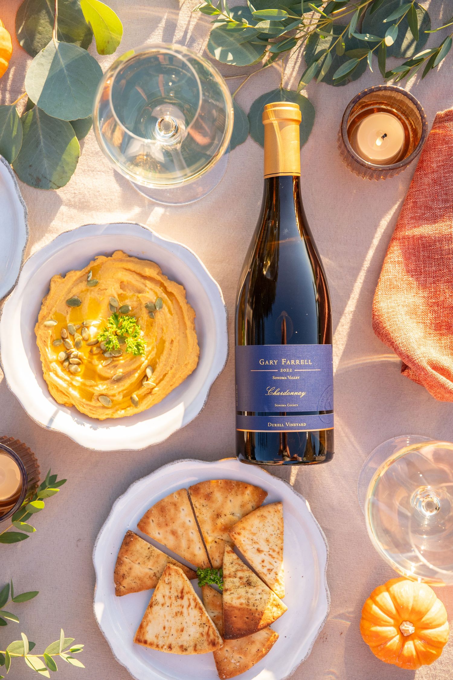 Overhead shot of hummus, pita bread, and wine bottles arranged on white plates with eucalyptus leaves.