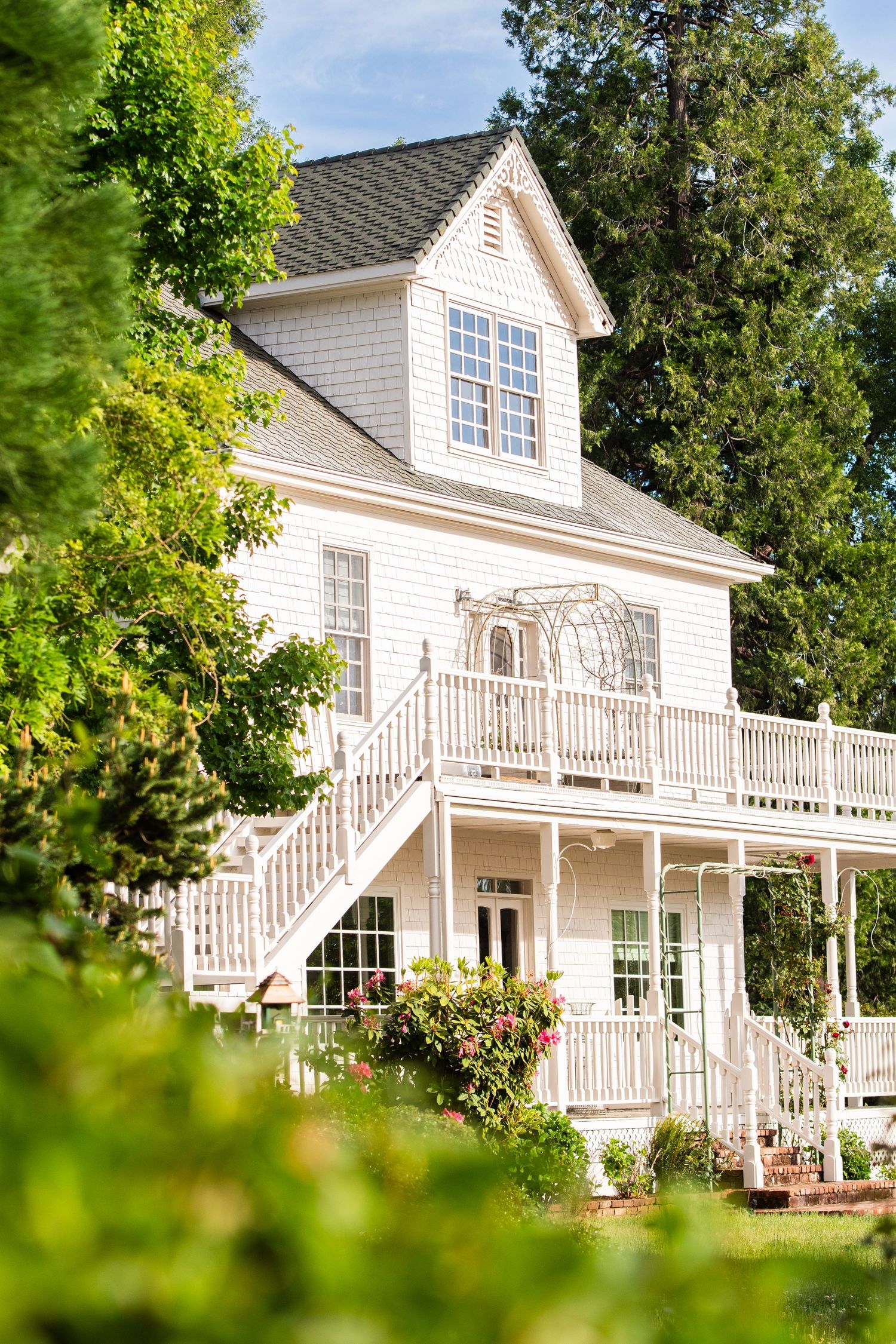 Elegant white cottage with dormer window and wrap-around porch surrounded by lush greenery.